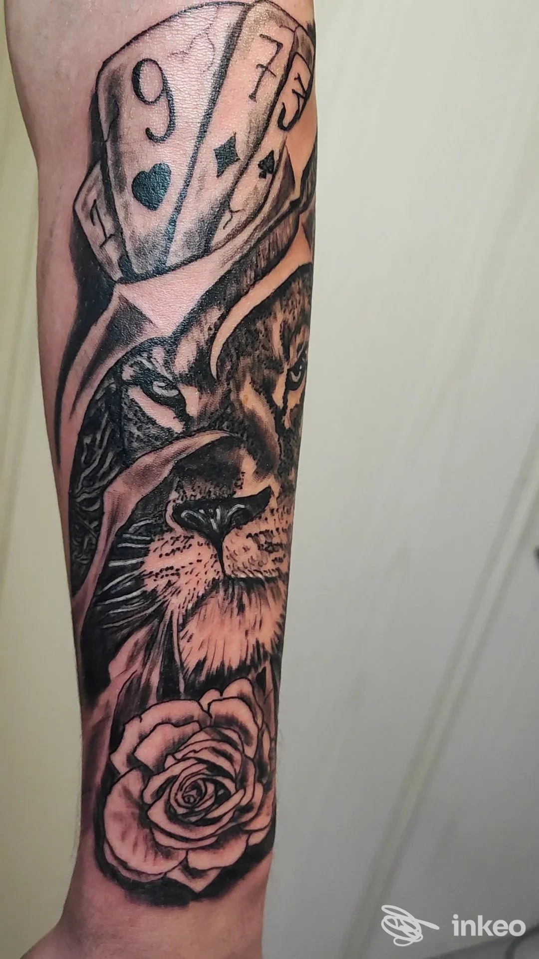 Tatuaggio sull'avambraccio con carte da gioco, leone e rosa in tonalità nero-grigio.