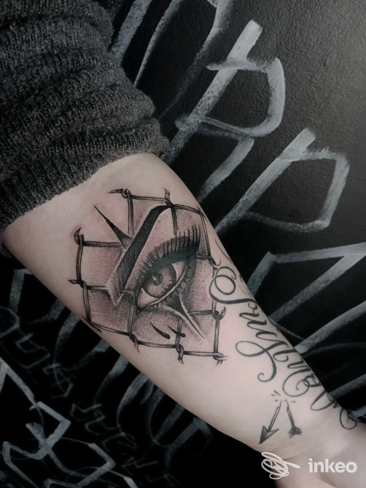 Primo piano di un tatuaggio sull'avambraccio con un occhio realistico incorniciato da punti di sutura e scritta decorativa, foto inviata da Malamore Tattoo, Massa.