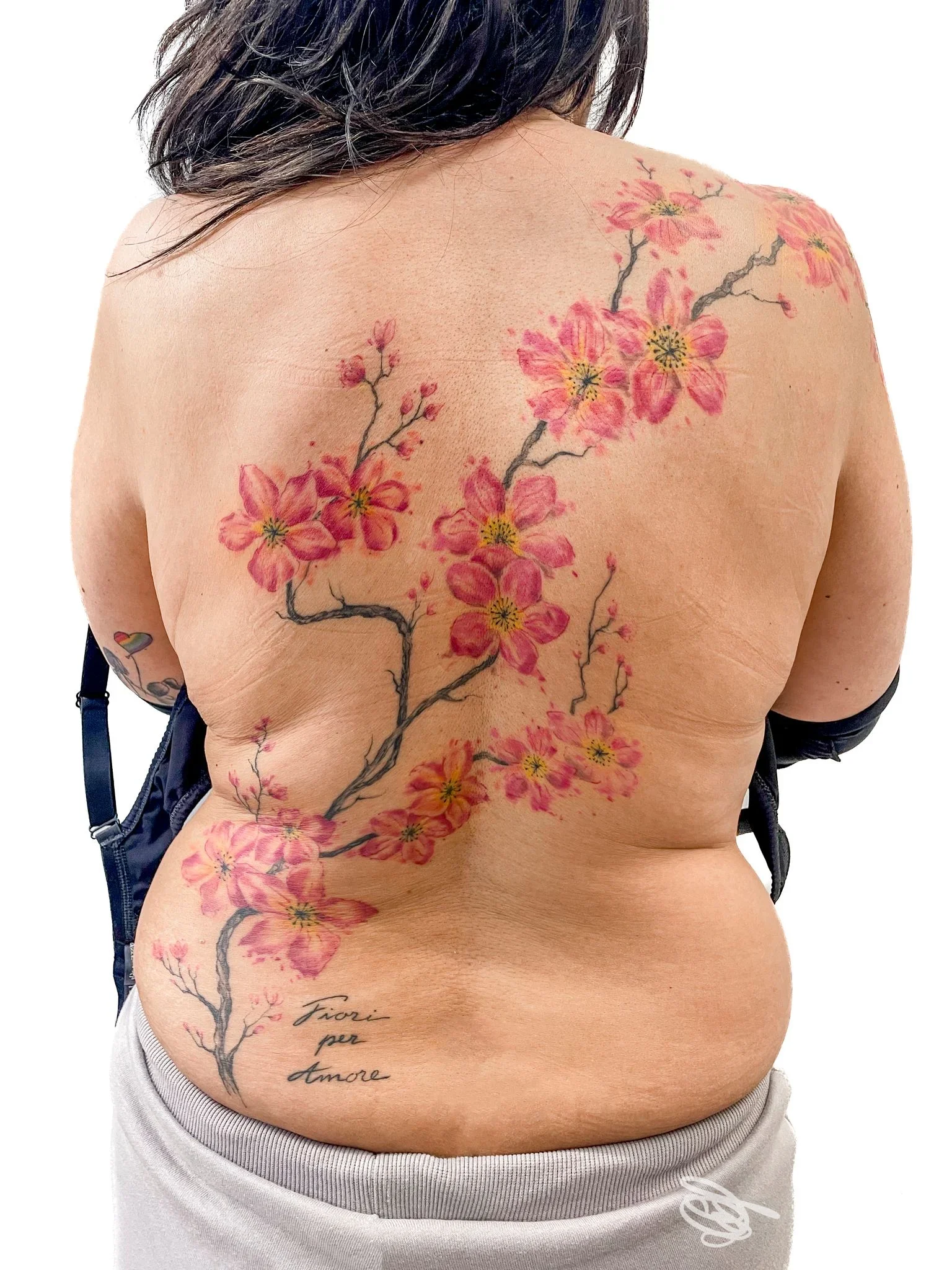 Grande tatuaggio floreale colorato che copre la schiena di una cliente, ramo di fiori rosa con stelo scuro e scritta in basso; immagine fornita da Mako Tattoo, Montignoso.