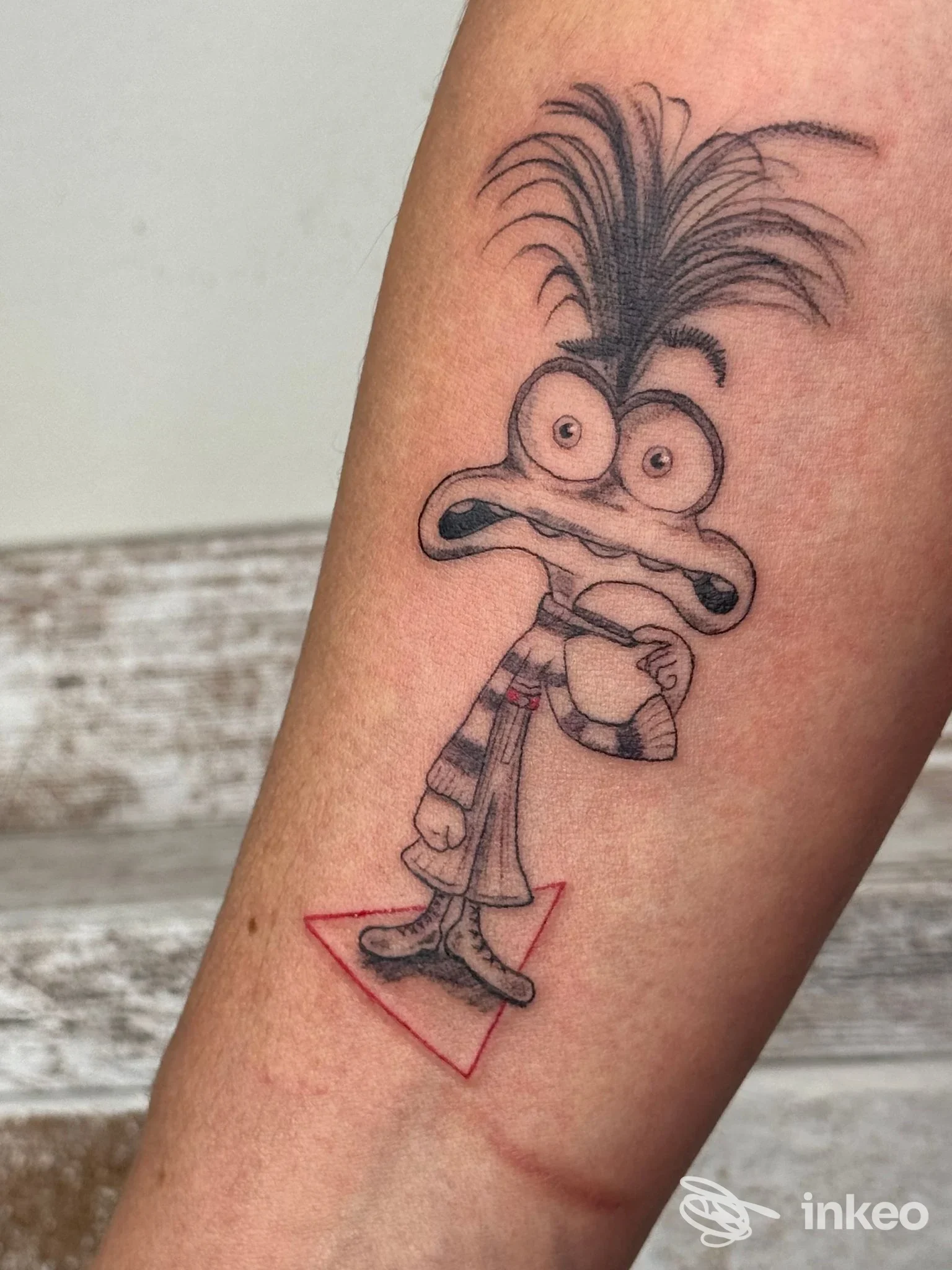 Tatuaggio su avambraccio di Nora Tattoo Studio, Scandicci: personaggio cartoonesco in nero con accento rosso su piccolo triangolo.