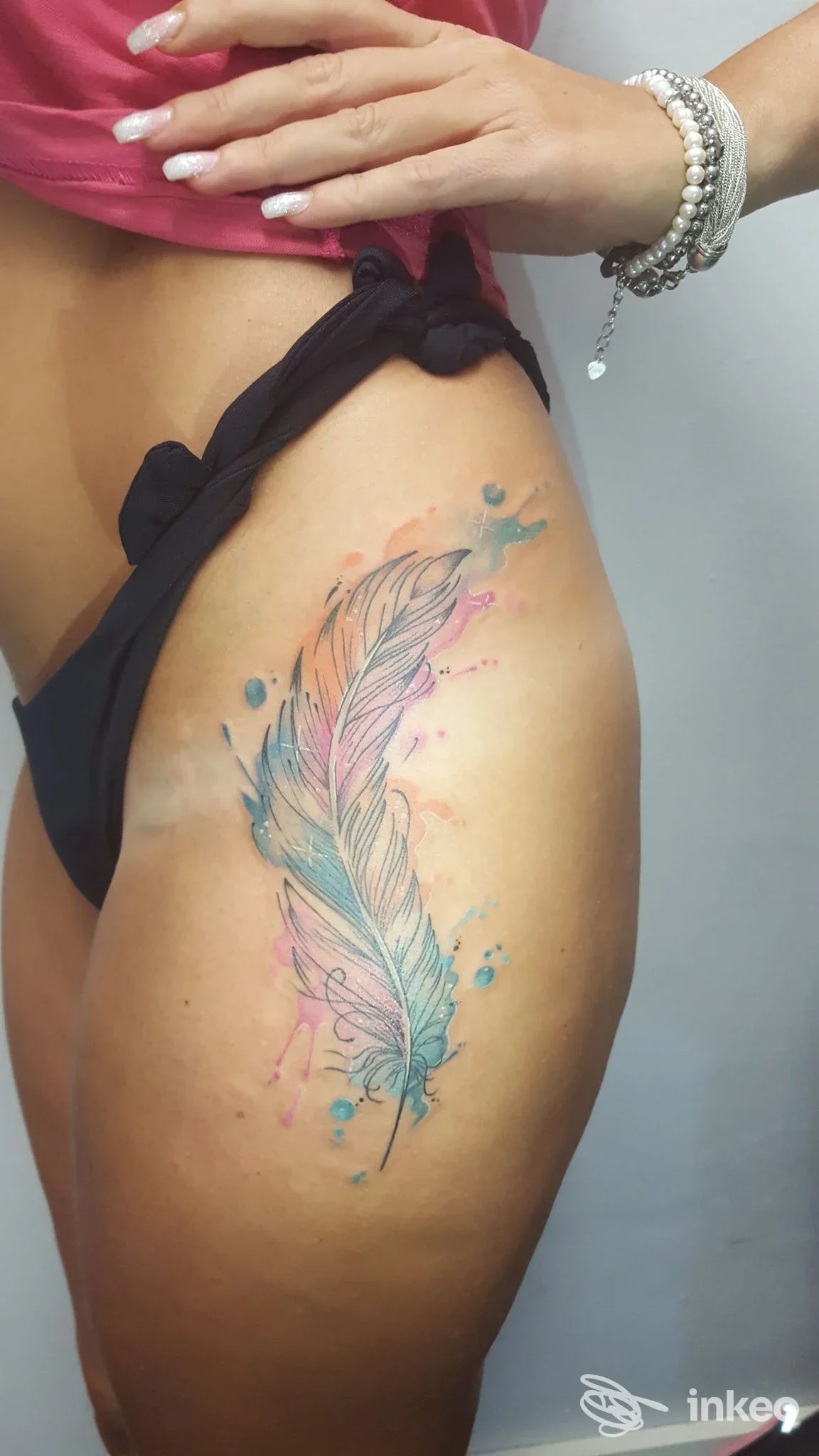Tatuaggio a piuma colorato su coscia/fianco femminile in stile acquerello, con sfumature rosa e azzurre. Tattoo Shop 669 Grosseto.