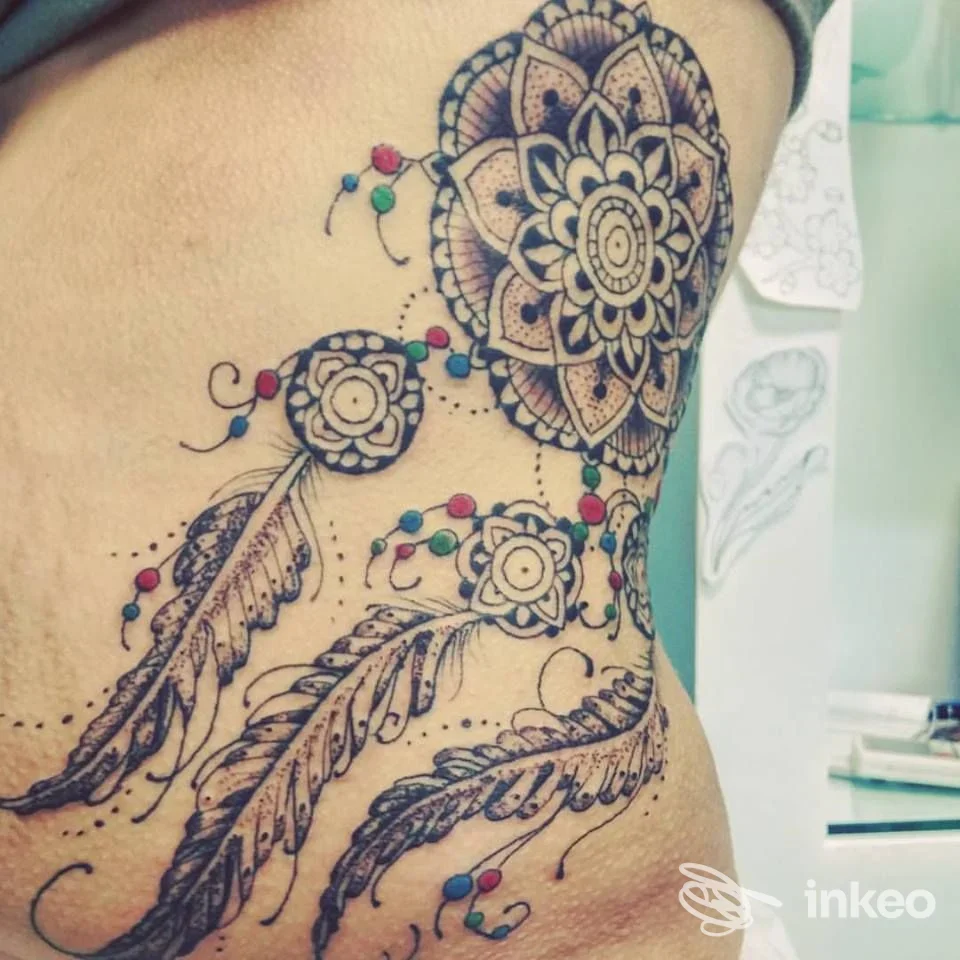 Tatuaggio ornamentale a mandala con piume e punti colorati, realizzato da Tatuando Di Squarcia Mara a Grosseto sul fianco.