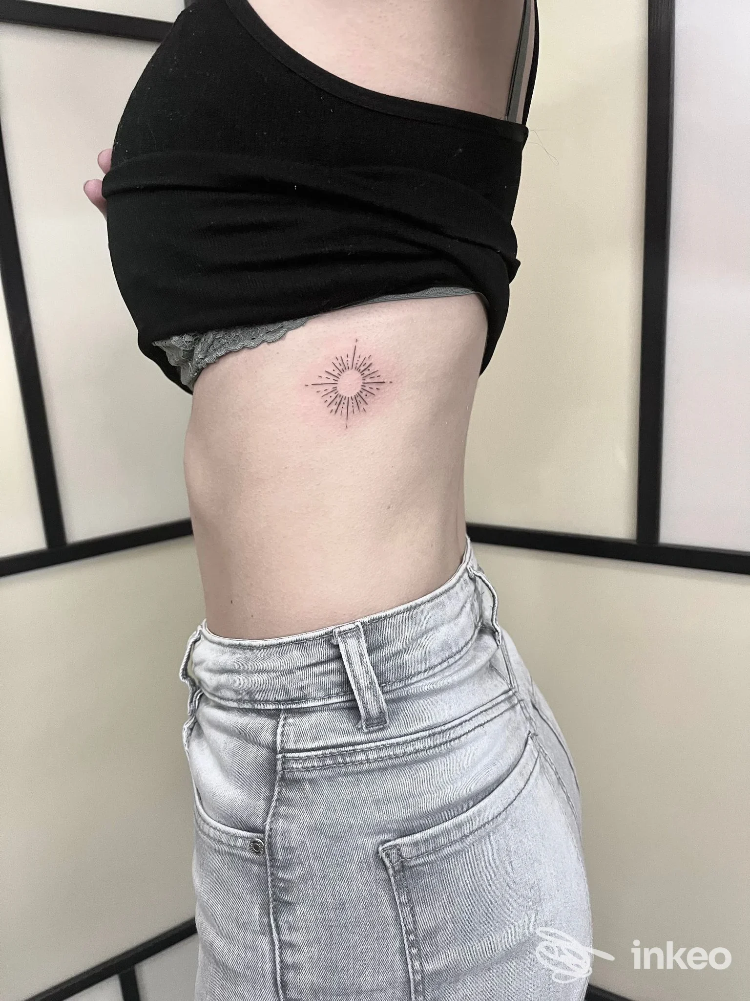 Piccolo tatuaggio a forma di sole sul fianco sinistro, disegno a linee sottili nere su pelle chiara, senza presenza di volto visibile.