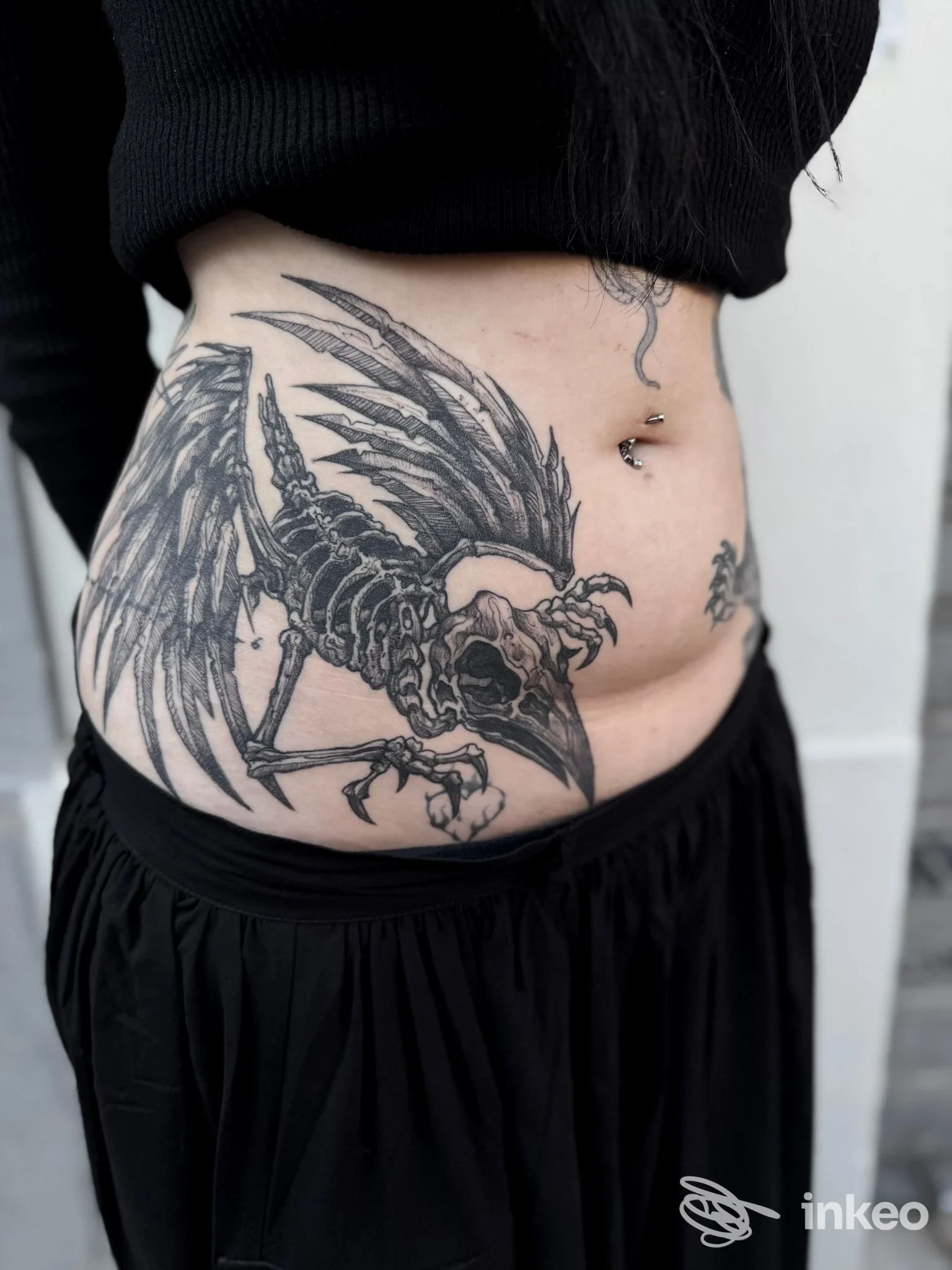 Dettaglio di addome con grande tatuaggio nero raffigurante uno scheletro/uccello e piercing all'ombelico. Foto per Adne Tattoo, Firenze.