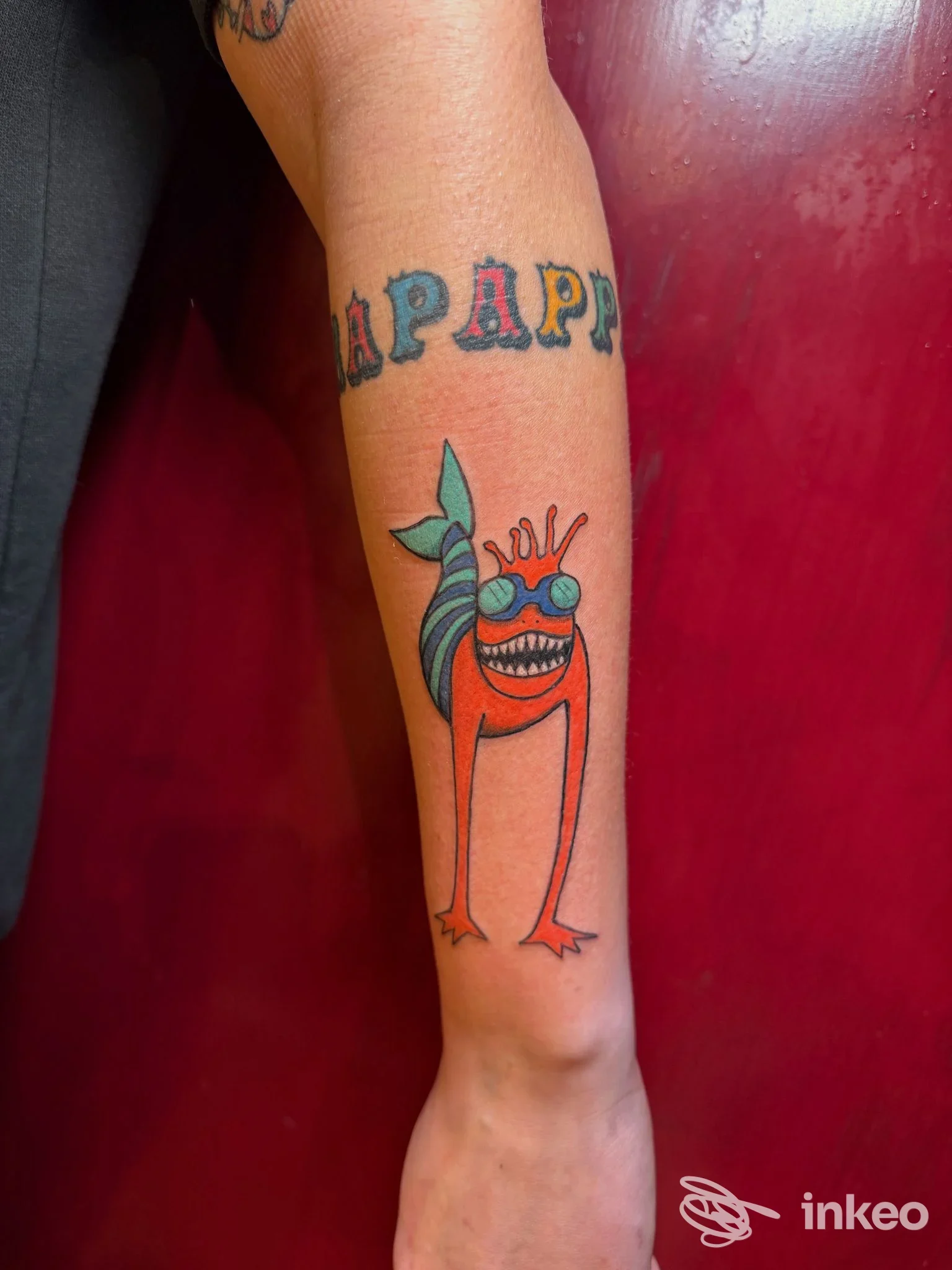Tatuaggio colorato su avambraccio con una creatura arancione dagli occhiali turchesi, foto fornita da Adne Tattoo, Firenze.