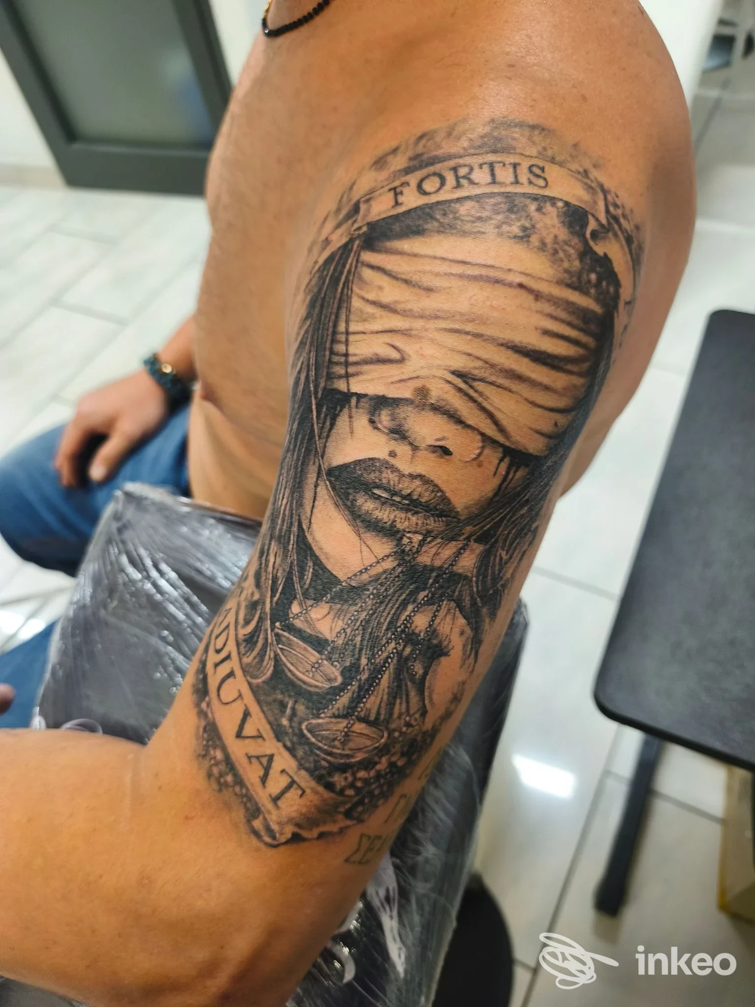 Tatuaggio realistico in bianco e nero sul braccio superiore: volto bendato e bilancia. VinzTattoo, Foiano della Chiana.