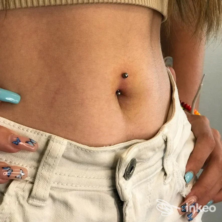Piercing all'ombelico su addome femminile con gioielli a due palline; mani con unghie decorate visibili ai bordi. Anele Piercing, Firenze.