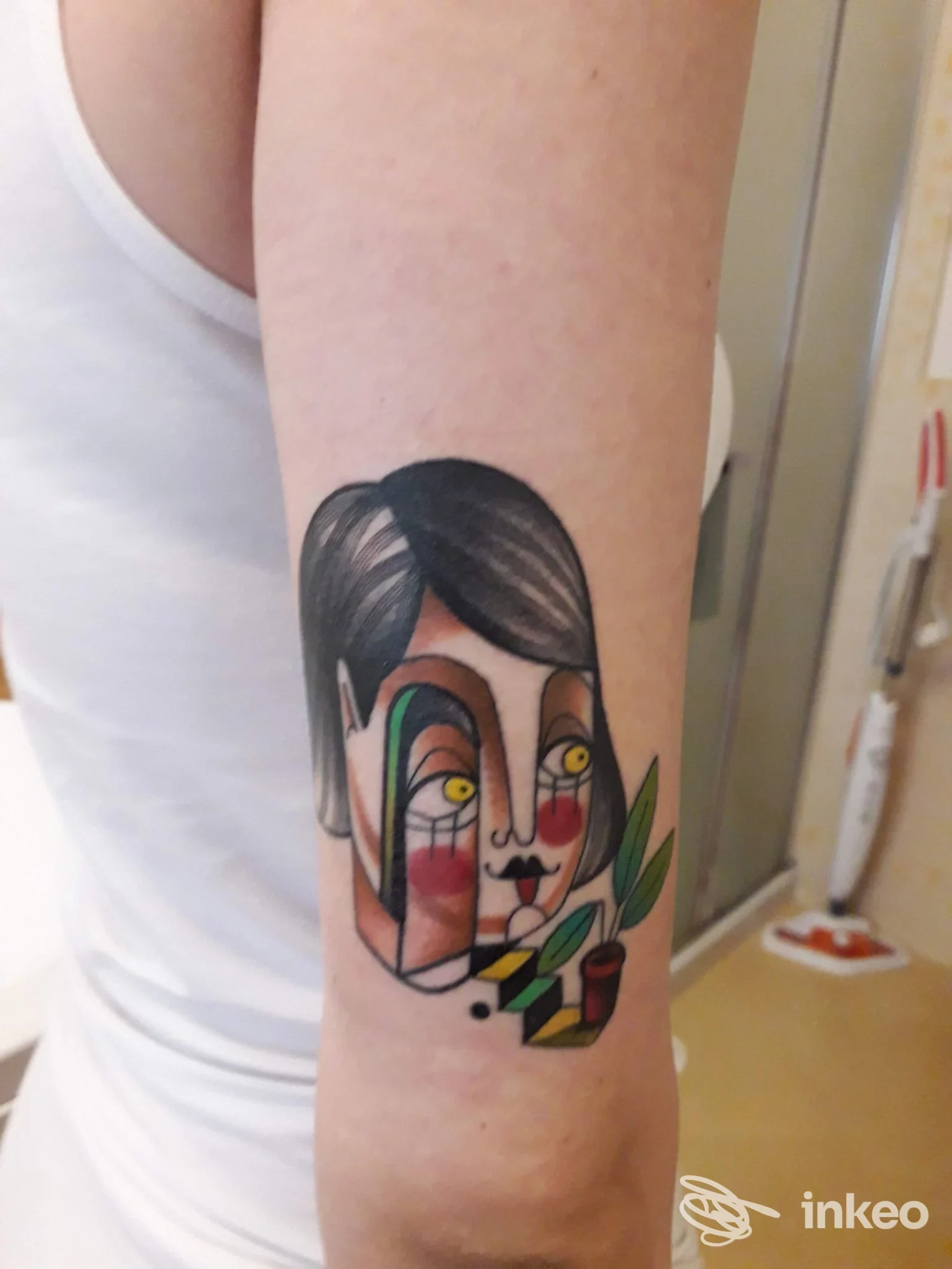 Tatuaggio sul braccio in stile astratto: volto stilizzato con colori vivaci e dettagli neri, eseguito sulla pelle dell’avambraccio dell’utente, con foglie verdi e accessori di colore.
