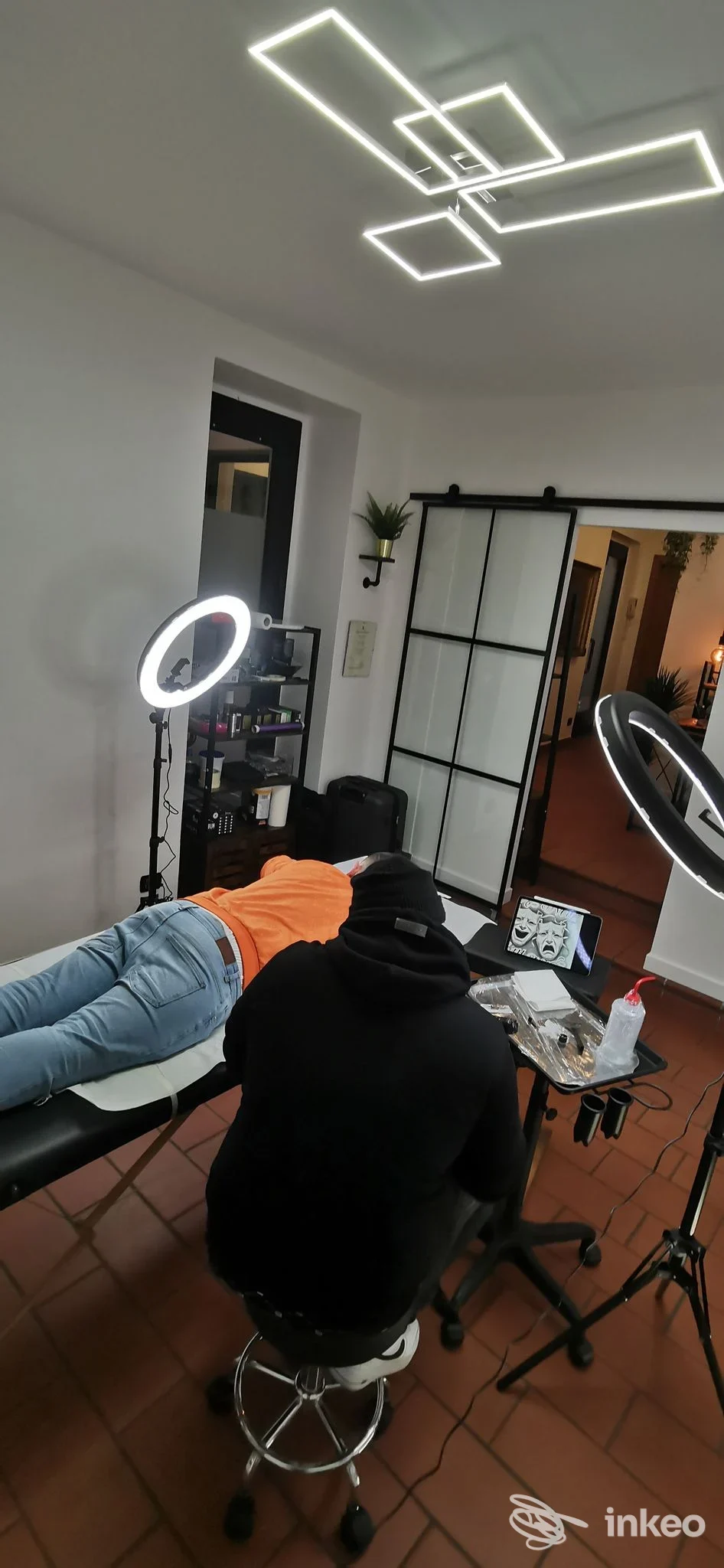 Interno dello studio Vibe Tattoo Atelier: una persona distesa su tavolo da tatuaggio mentre l'artista lavora sulla pelle; luci a LED e ring light, ambiente moderno.