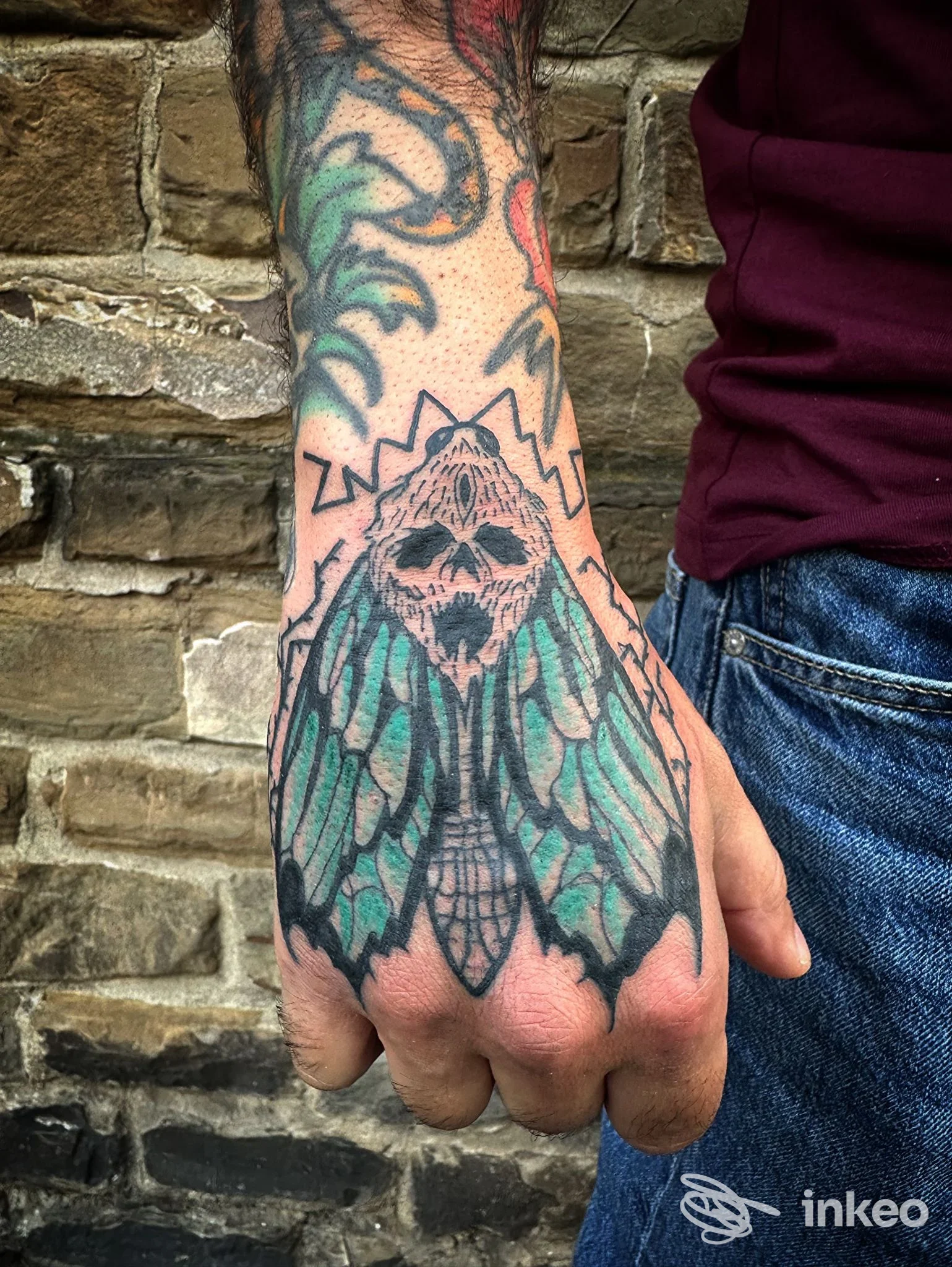 Tatuaggio sul dorso della mano: falena stilizzata con teschio centrale e colori verdi/turchesi su sfondo muro; Black Mamba Tattoo, Firenze.