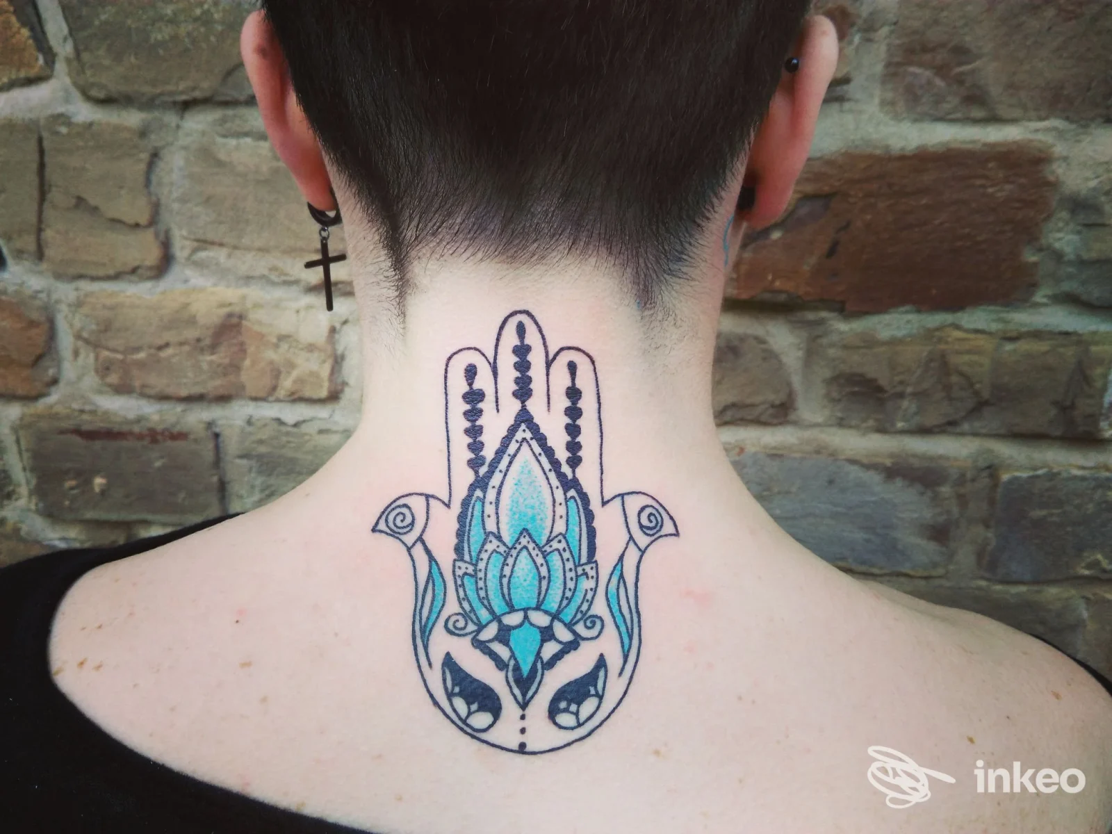 Tatuaggio a mano di Fatima (hamsa) colorata sulla nuca di una persona, eseguito da Black Mamba Tattoo a Firenze, su sfondo muro di mattoni.