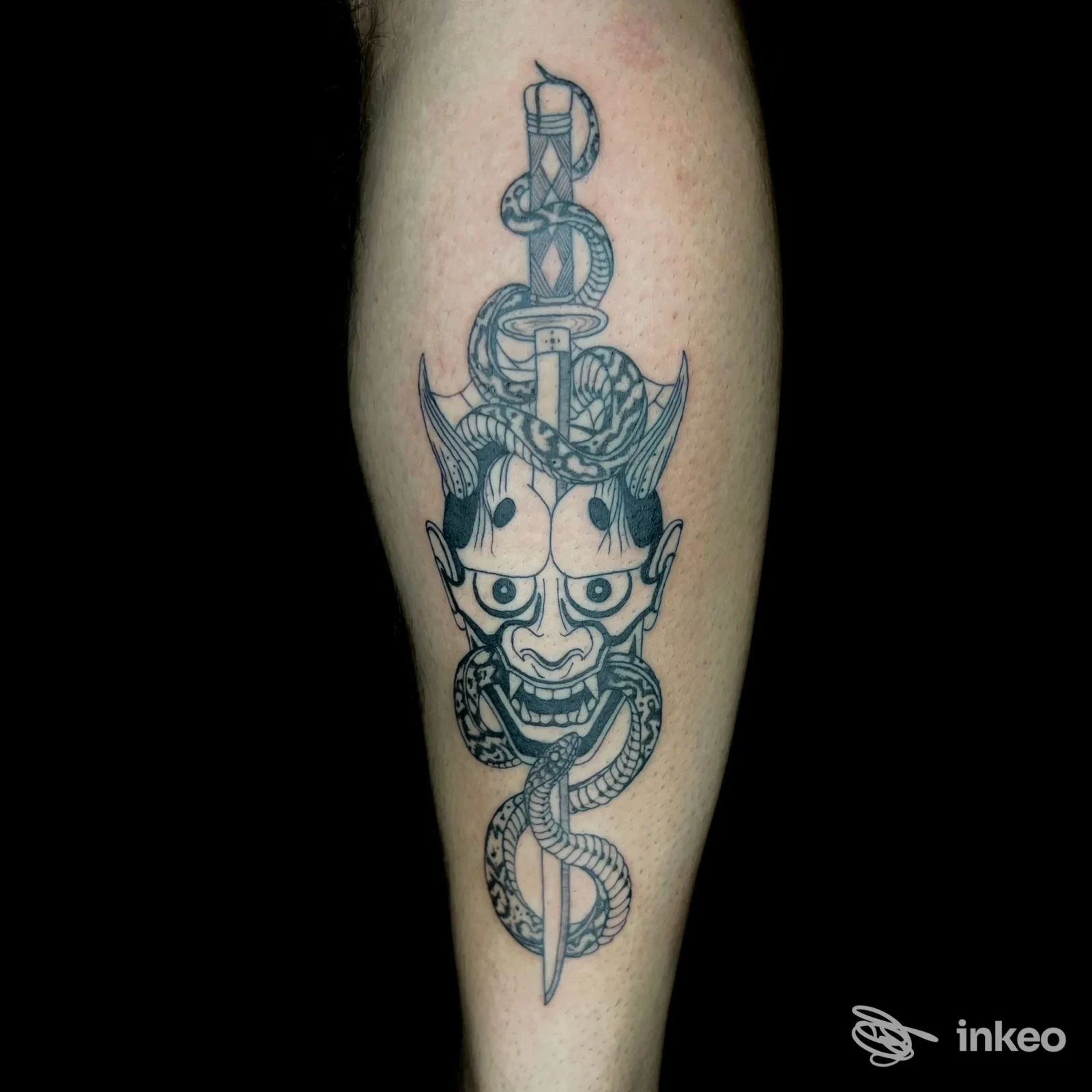 Tatuaggio su polpaccio con maschera demoniaca e serpente avvolto attorno a un pugnale, pelle chiara su sfondo nero.
