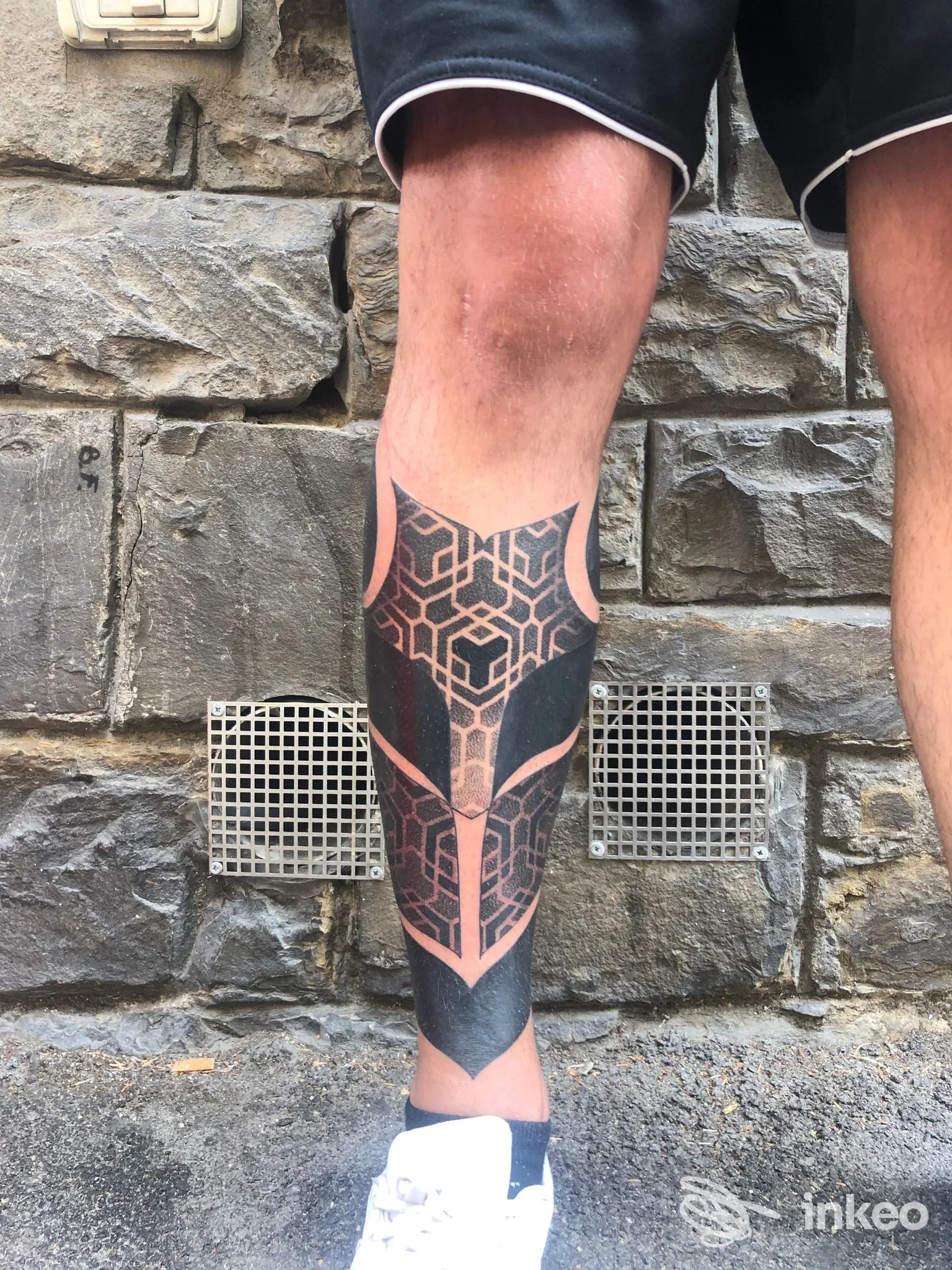 Tatuaggio geometrico nero sul polpaccio di una persona, scatto verticale davanti a un muro in pietra. BLACK FLOW Tattoo Studio, Firenze.