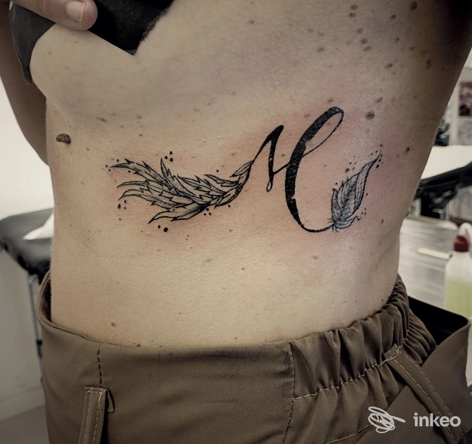 Tatuaggio a inchiostro nero sul fianco con una M stilizzata e piume, foto dallo studio Chimera Tattoo Studio a Firenze.