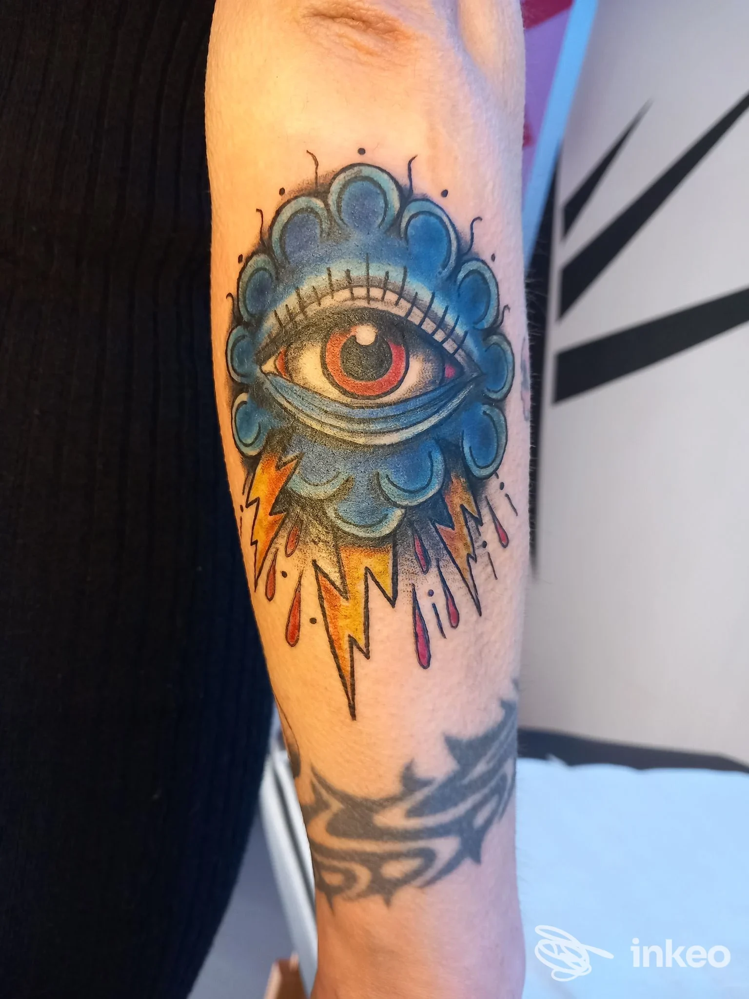 Tatuaggio colorato su avambraccio con grande occhio stilizzato racchiuso in una nuvola blu e fulmini; immagine per CERBERUS TATTOO SHOP ®️ a La Rosa.