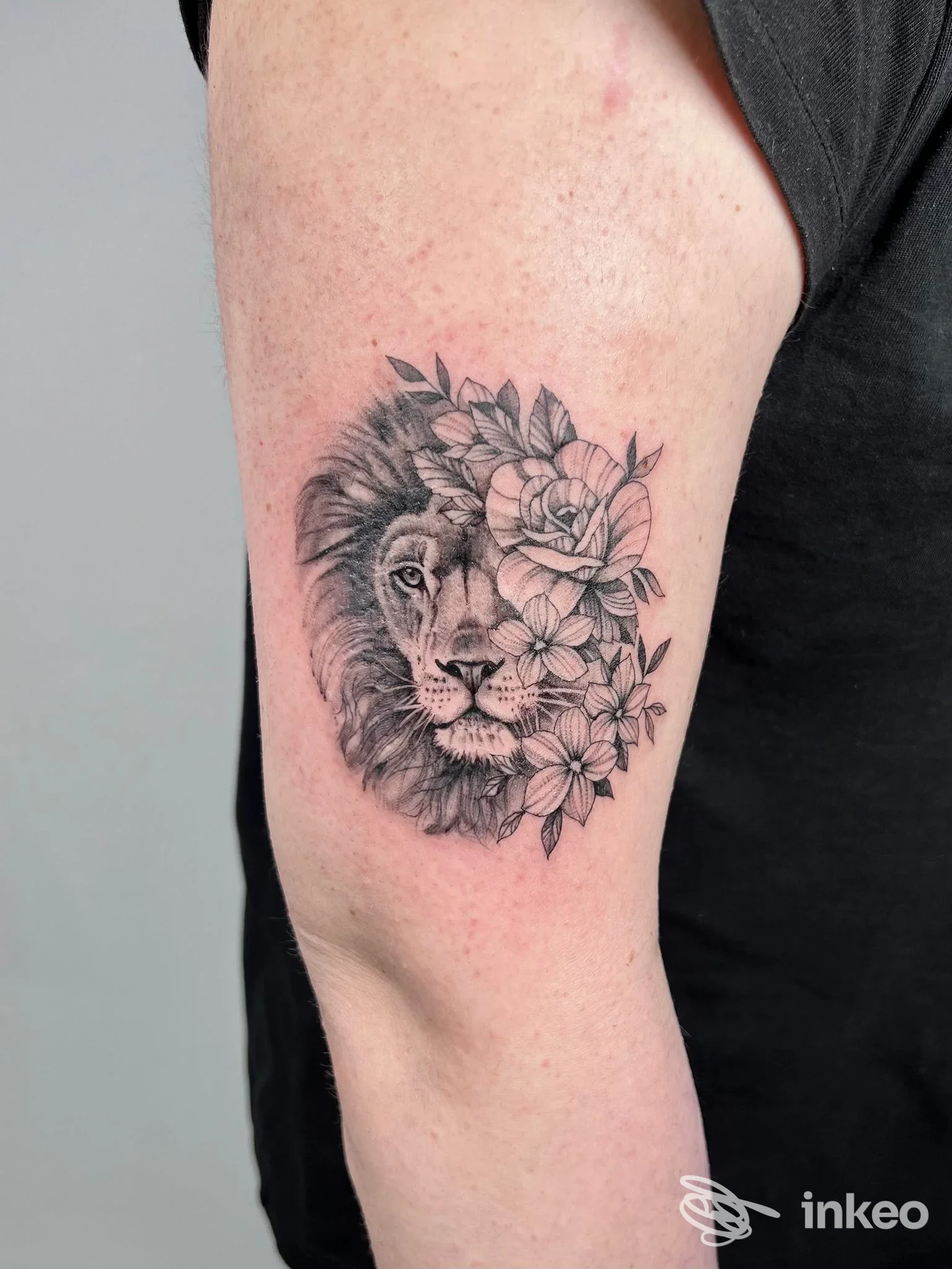 Tatuaggio realistico in bianco e nero di leone parzialmente circondato da fiori sul braccio superiore. Efi Artist Tattoo&Jewels, Montecatini Terme.
