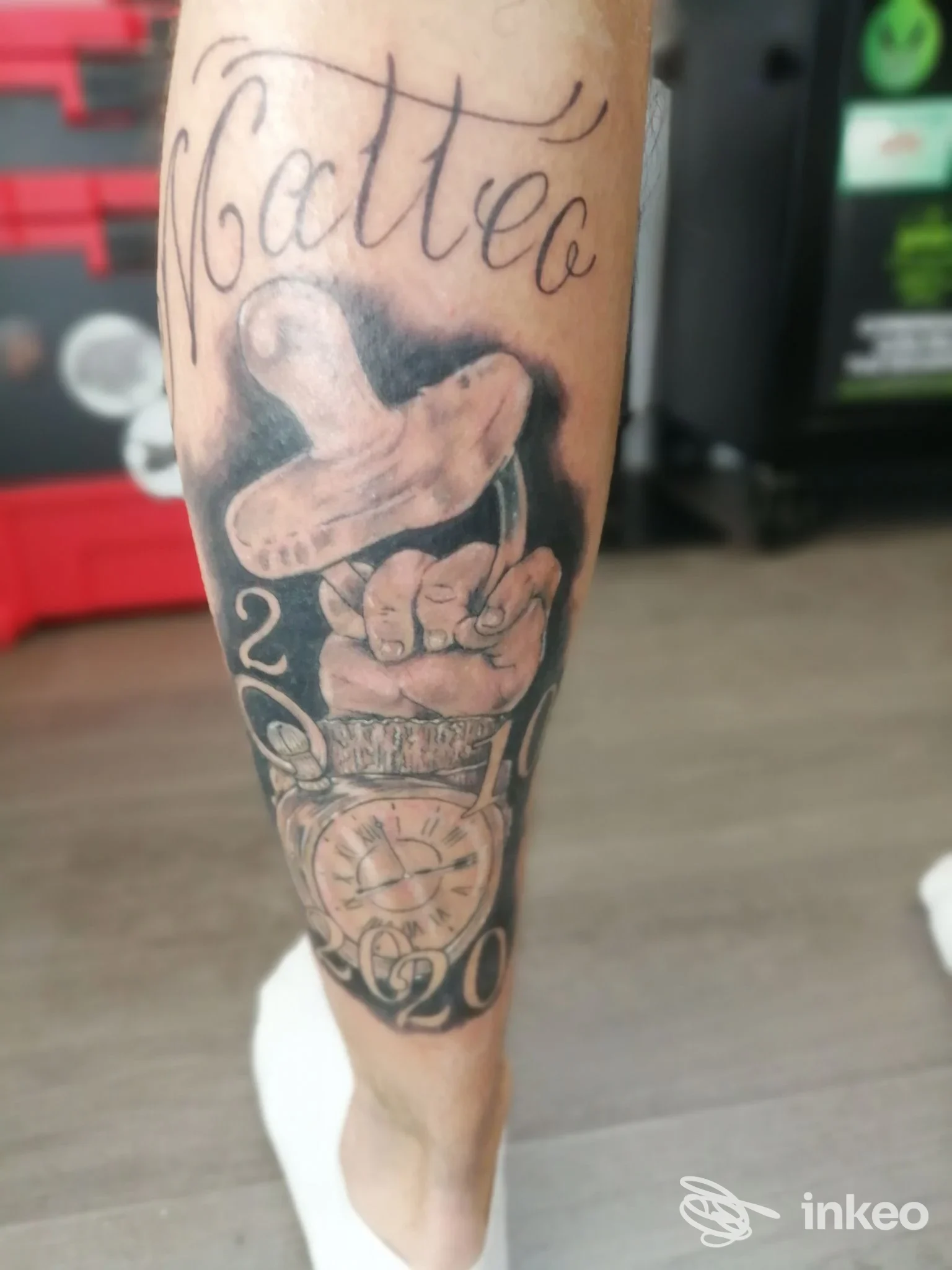 Tatuaggio black & grey sul polpaccio con la scritta "Matteo", una mano che regge un oggetto e un orologio da tasca. Inkrinato Tattoo Studio, Pisa.