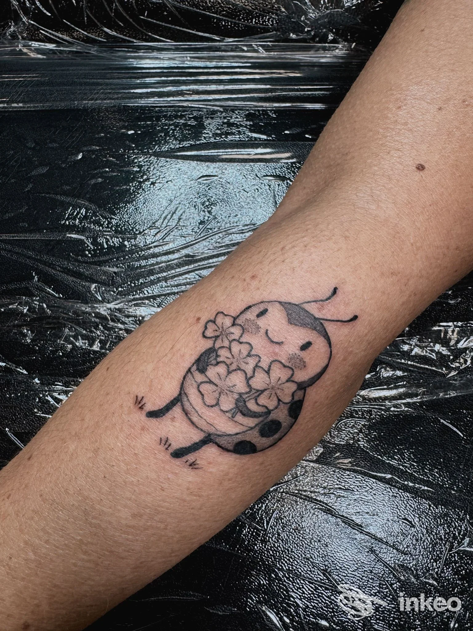 Tatuaggio su avambraccio di una coccinella stilizzata che tiene fiori, linee nere e sfumature grigie. Scatto per Inlè Tattoo Studio, Lucca.