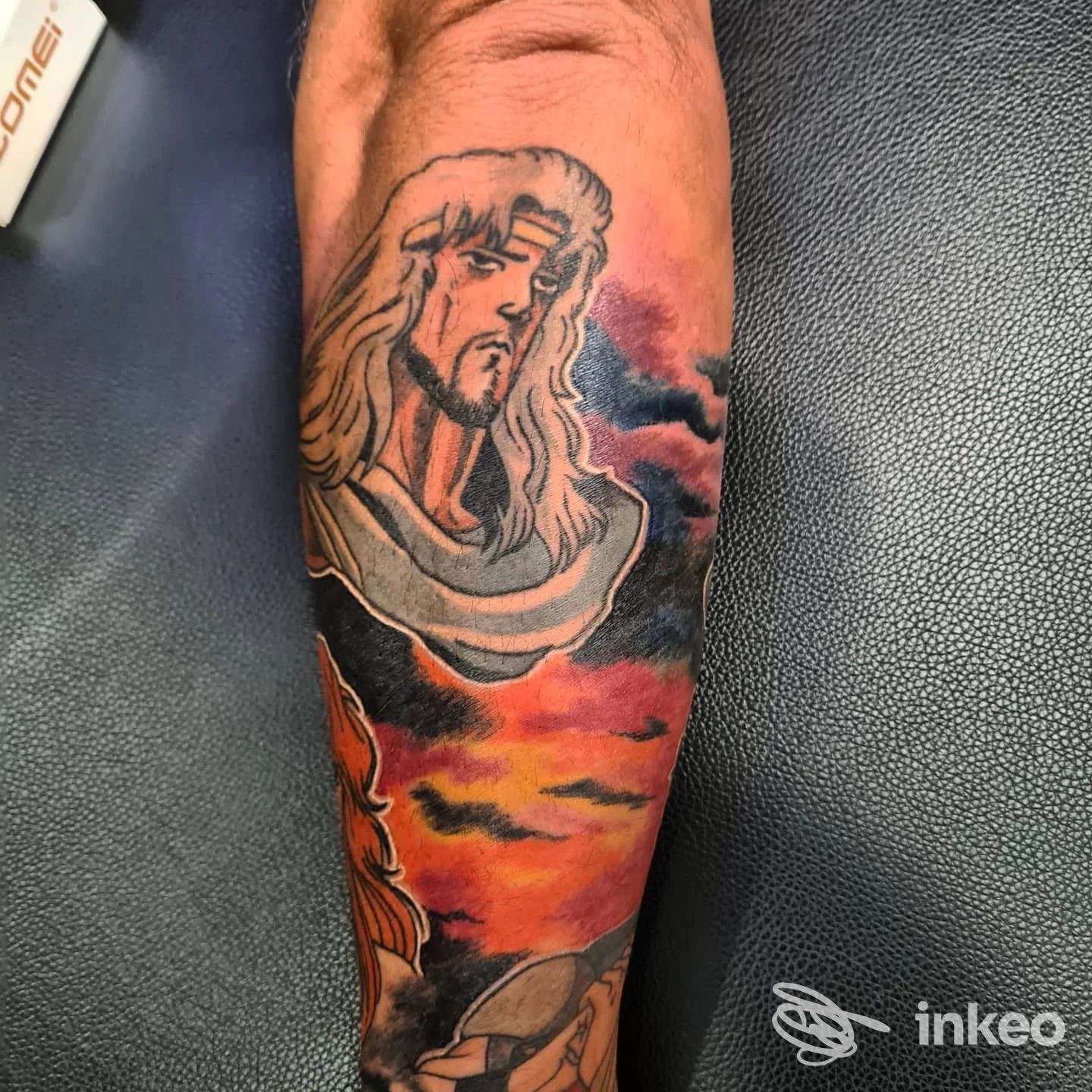Avambraccio con tatuaggio colorato raffigurante volto sacro e cielo infuocato, Love Hate Tattoo Studio, Marina di Carrara.
