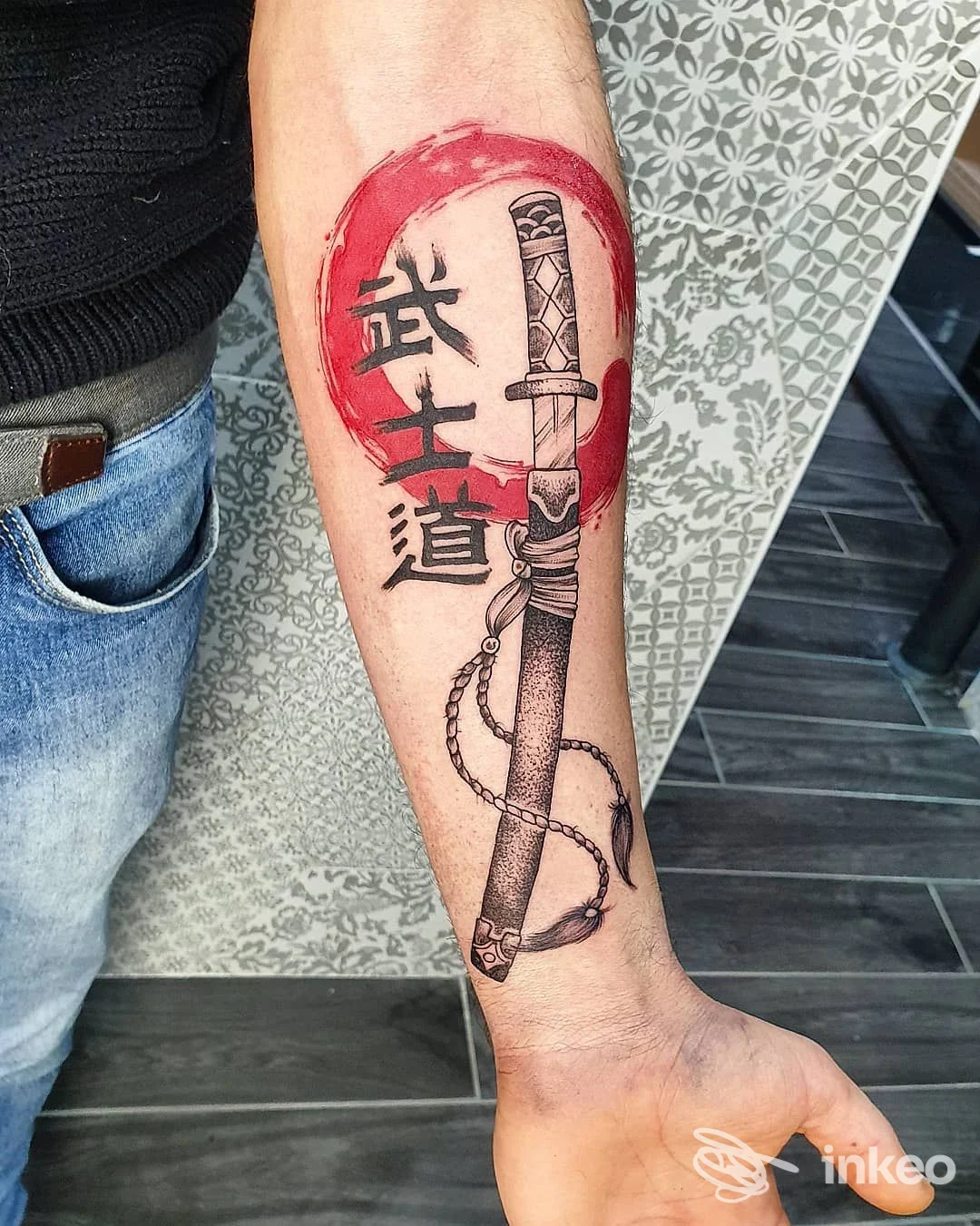 Tatuaggio sull'avambraccio sinistro raffigurante una katana con dettagli puntinati, corde e ideogrammi giapponesi dentro un cerchio rosso, realizzato da Madame Black Tattoo a Livorno.