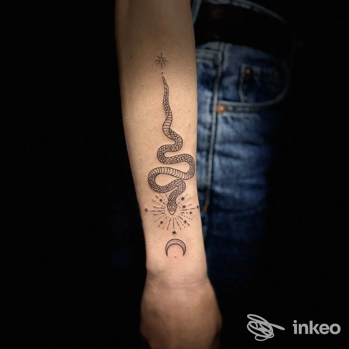 Tattoo visibile sull’avambraccio di una persona: serpente avvolto, decorazioni stellate e mezzaluna, sfondo scuro.