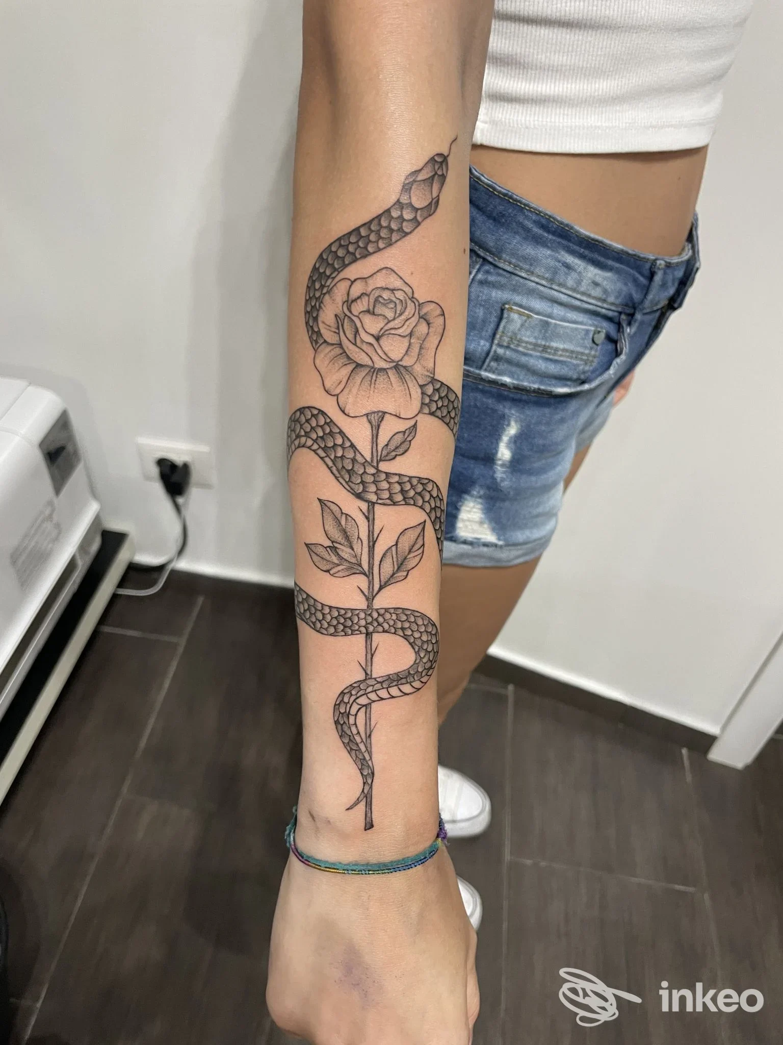 Tattoo su avambraccio con serpente avvolgente e rosa al centro, disegno in bianco e nero su pelle, stile linework, braccio di donna rurata.