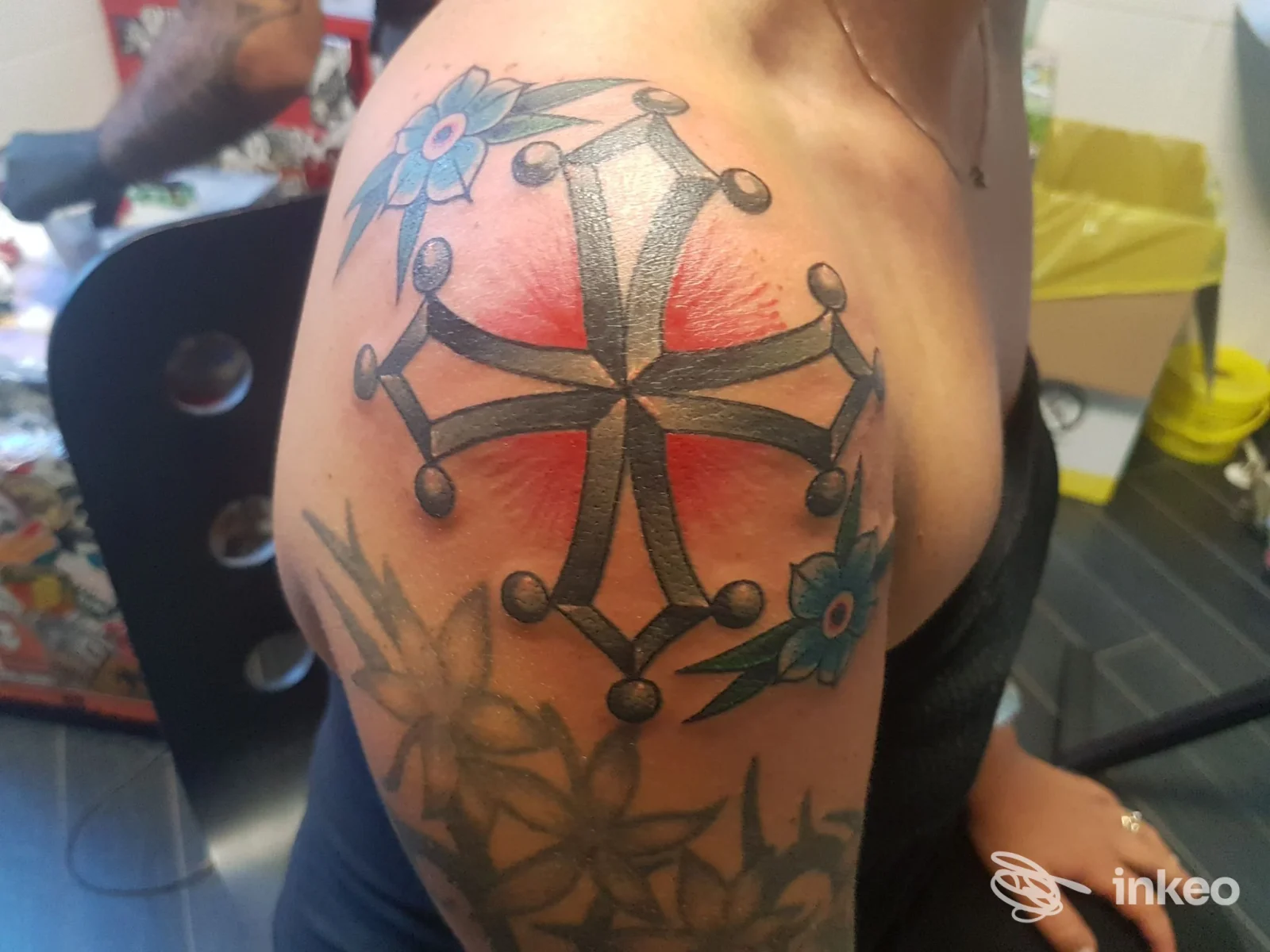 Tatuaggio a croce con sfumatura rossa e fiori blu sulla spalla, foto fornita da Scripta Manent Tattoo Studio, Pisa.