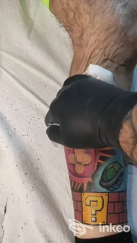 Avambraccio con tatuaggio colorato a tema videogame (blocco '?', cerotto illustrato), mano del tatuatore con guanto nero. Scripta Manent Tattoo Studio, Pisa.