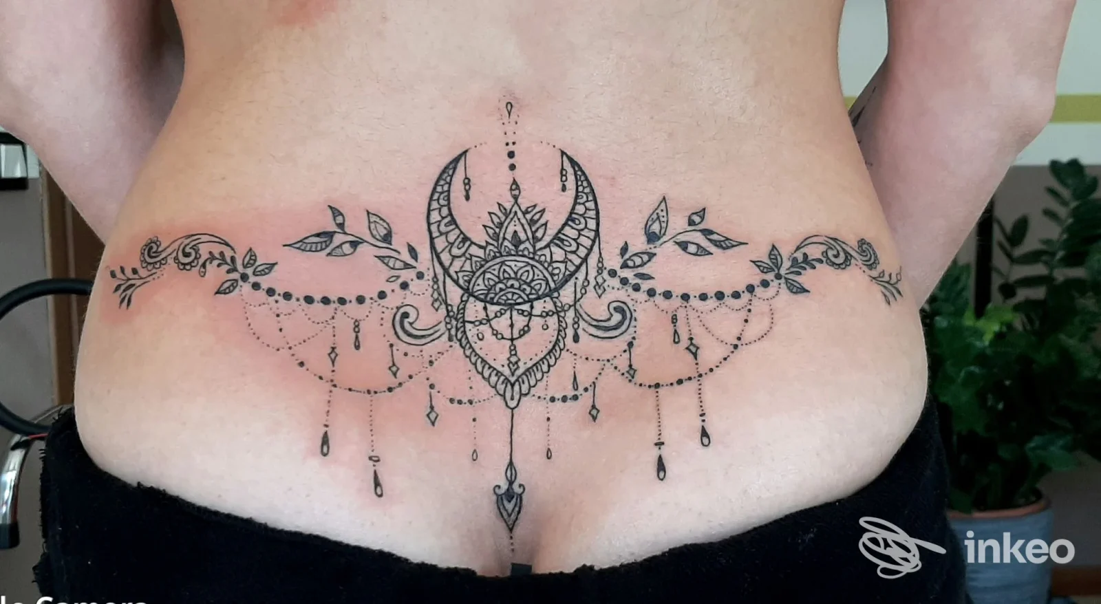 Tatuaggio sul fianco posteriore con motivo ornato e luna crescente al centro, con pendenti, realizzato sulla pelle.