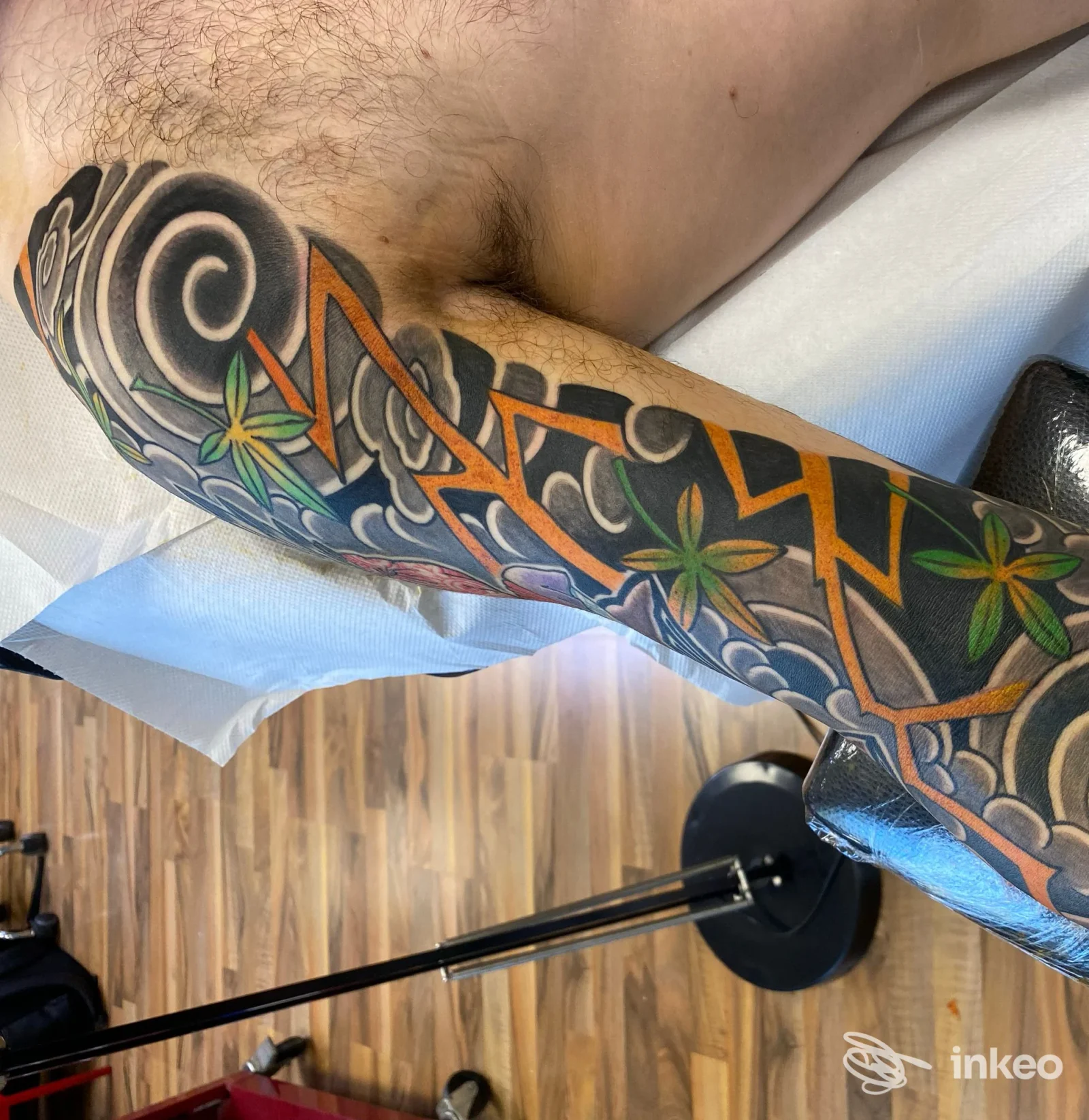 Tatuaggio a manica su braccio maschile con motivi neri, arancio e fiori verdi, foto fornita da Tattooing With Love, Monsummano Terme.