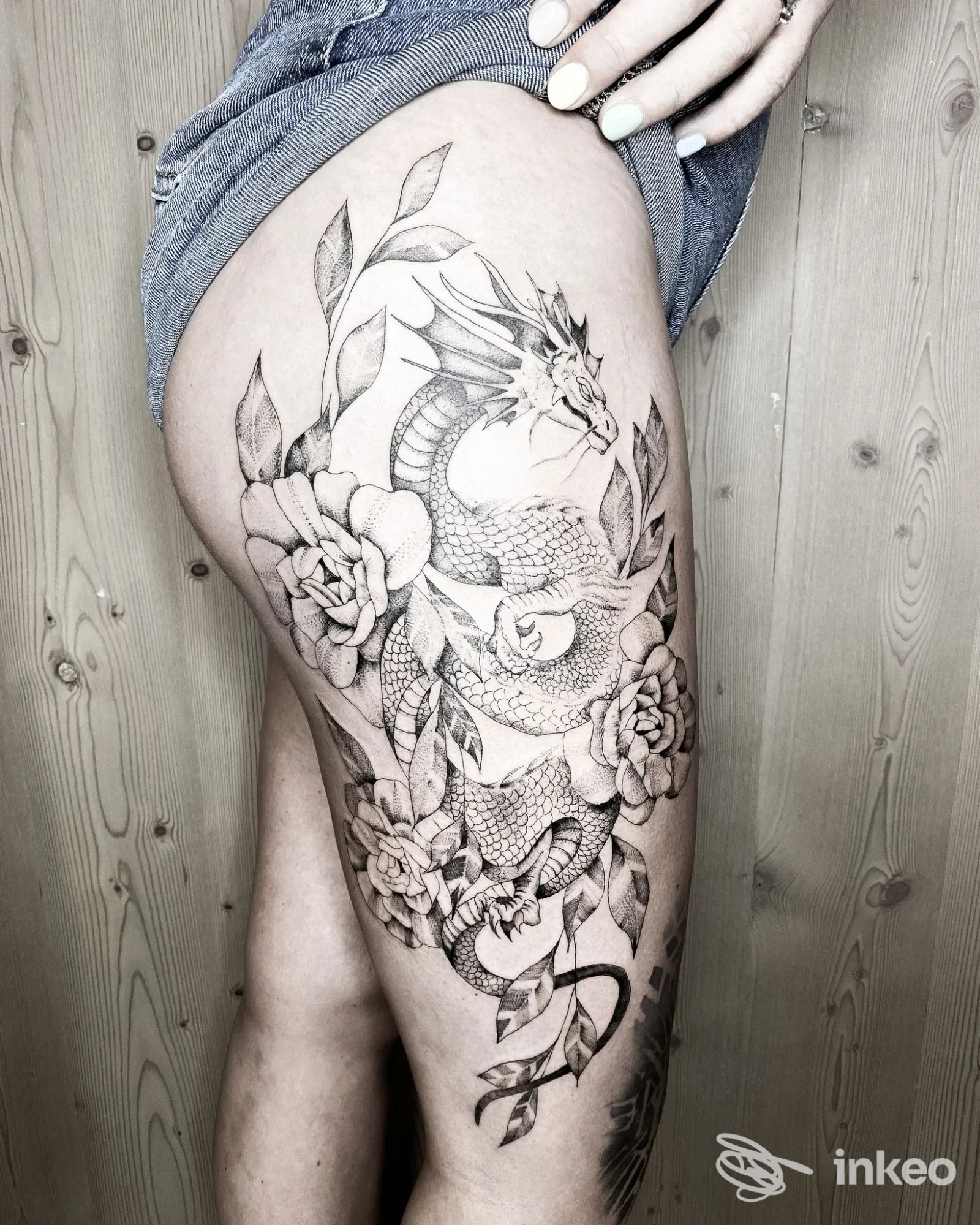 Tatuaggio dettagliato di drago avvolto tra rose e foglie sulla coscia, realizzato da Athena Tattoo Studio a Cles.