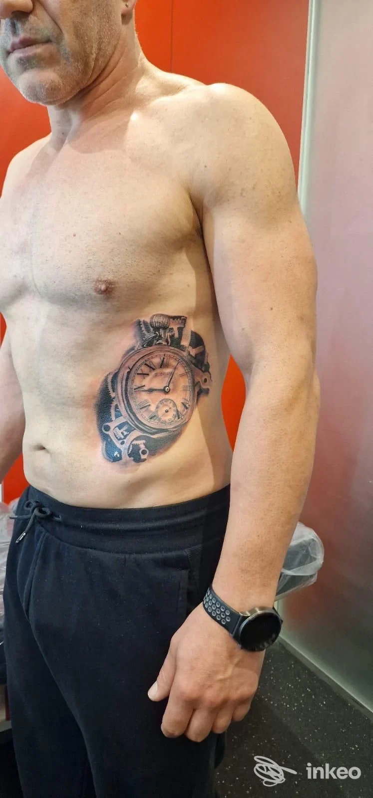 Uomo in piedi che mostra un tatuaggio realistico di orologio sul fianco destro, foto dallo studio Blackpanther Piercing & Tattoo Di Alessandro Mancin, Trento.