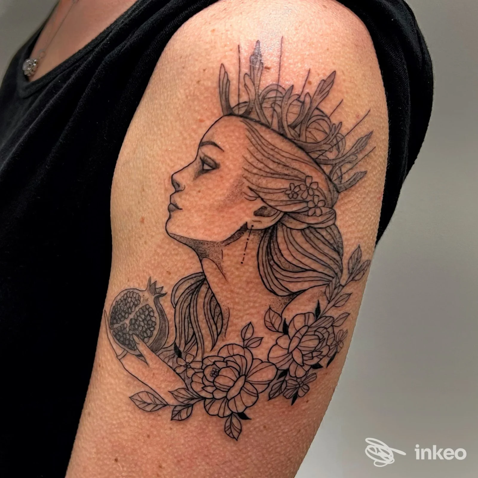 Tatuaggio linework a profilo femminile con melograno e fiori sul braccio superiore, realizzato da DEVI TATTOO a Levico Terme.