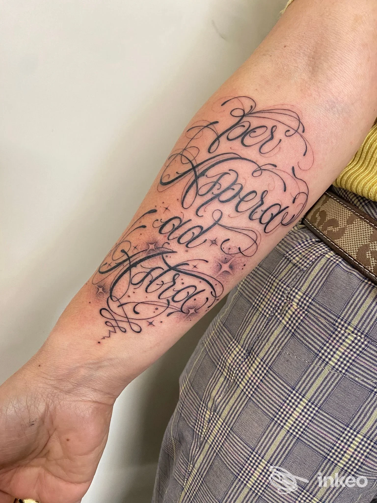 Tatuaggio scriptato ornamentale sull'avambraccio sinistro: parole in corsivo con riccioli e piccoli elementi decorativi, nessun volto visibile.