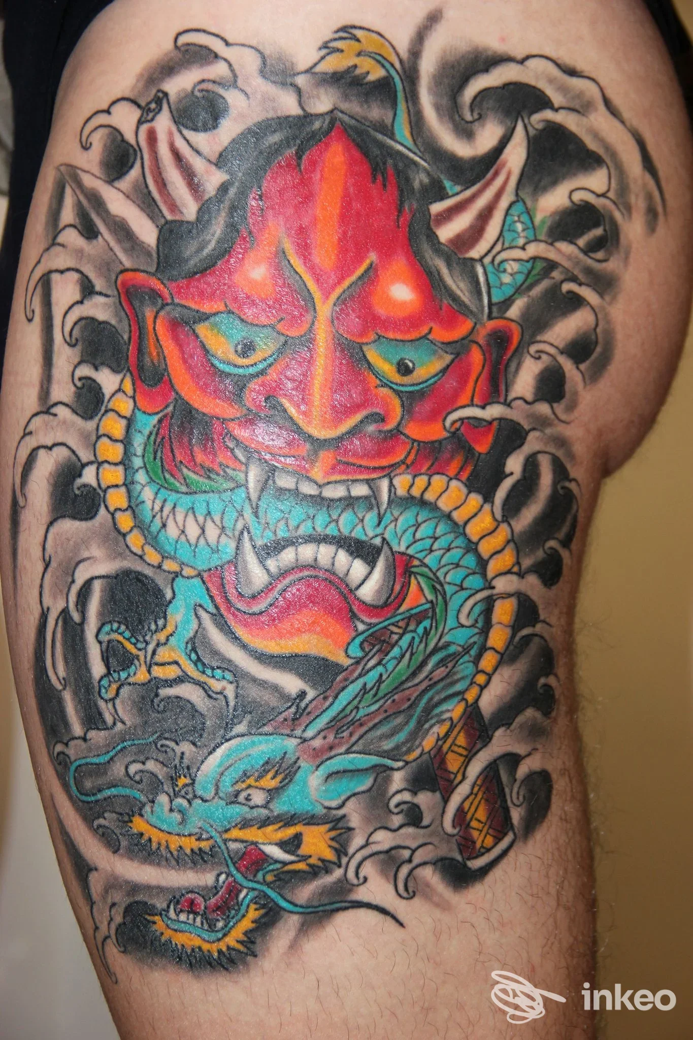 Grande tatuaggio a colori su coscia che ritrae una maschera demonica rossa e un drago turchese, foto di Red Dragon Tattoo a Pellizzano.