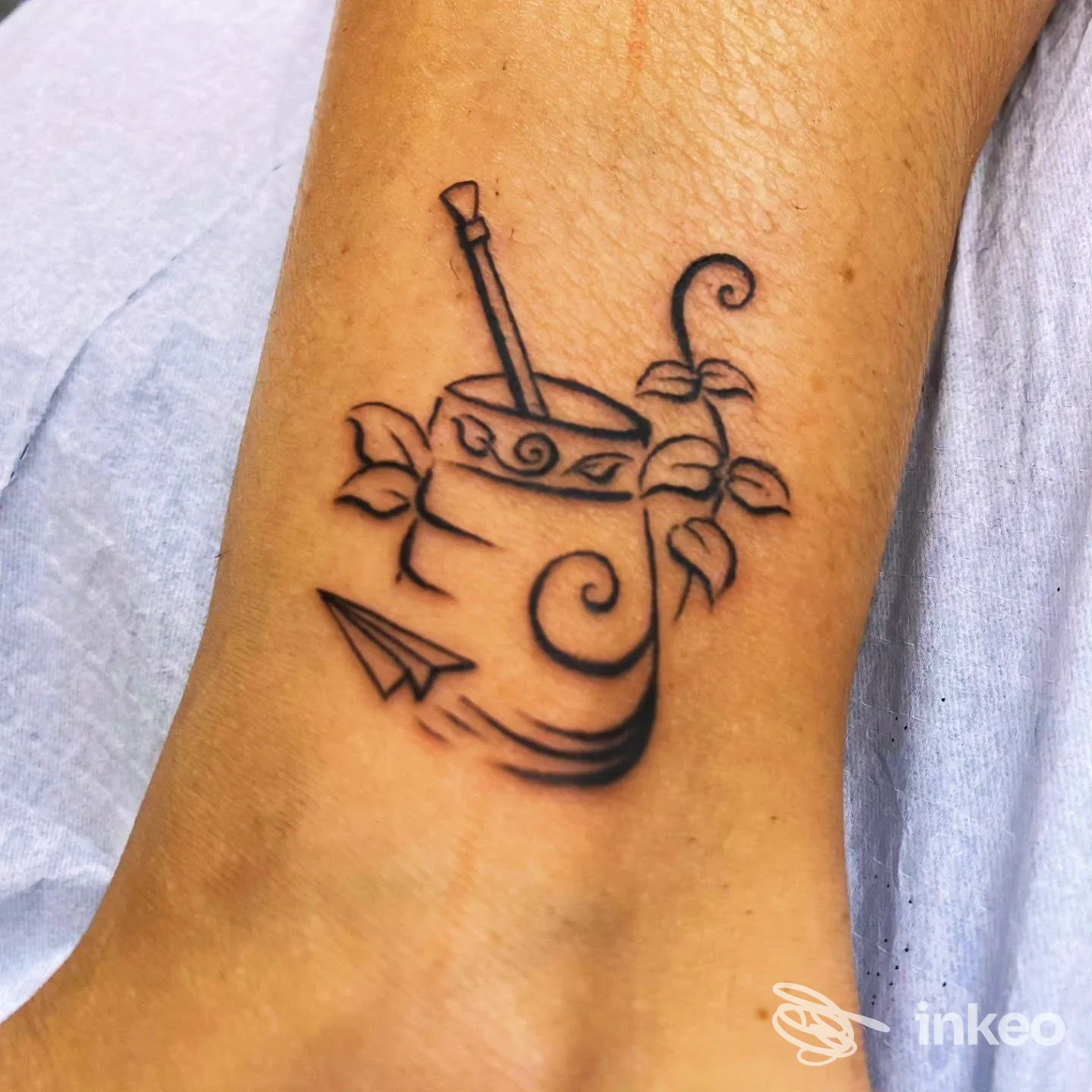 Tatuaggio minimal a linee nere vicino alla caviglia: tazza con bastoncino, foglie e un piccolo aeroplanino. Shakende Tattoo Studio, Fai della Paganella.