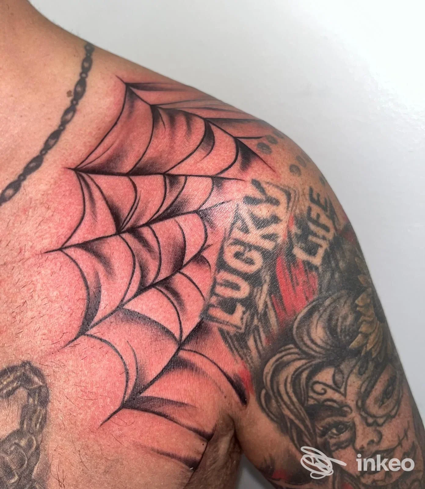 Tatuaggi sulla spalla e parte superiore del petto, ragnatela e volto stilizzato; Tattoo Fatline, Bolzano.