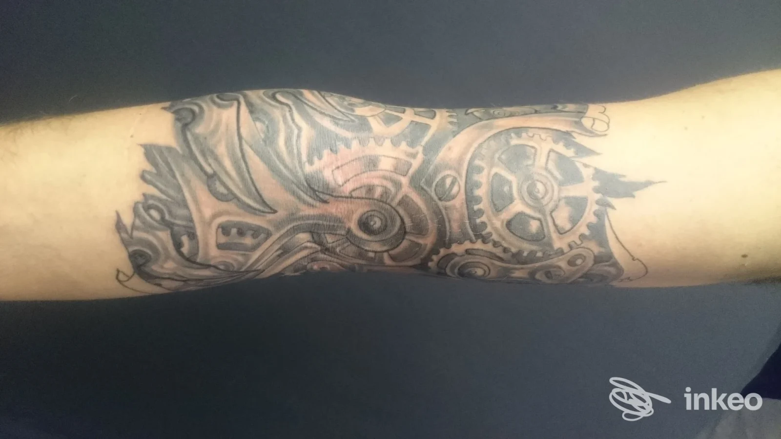 Avambraccio con tatuaggio in stile meccanico, trattato in chiaroscuro nero. Non si vede alcun volto, solo la pelle con il disegno.