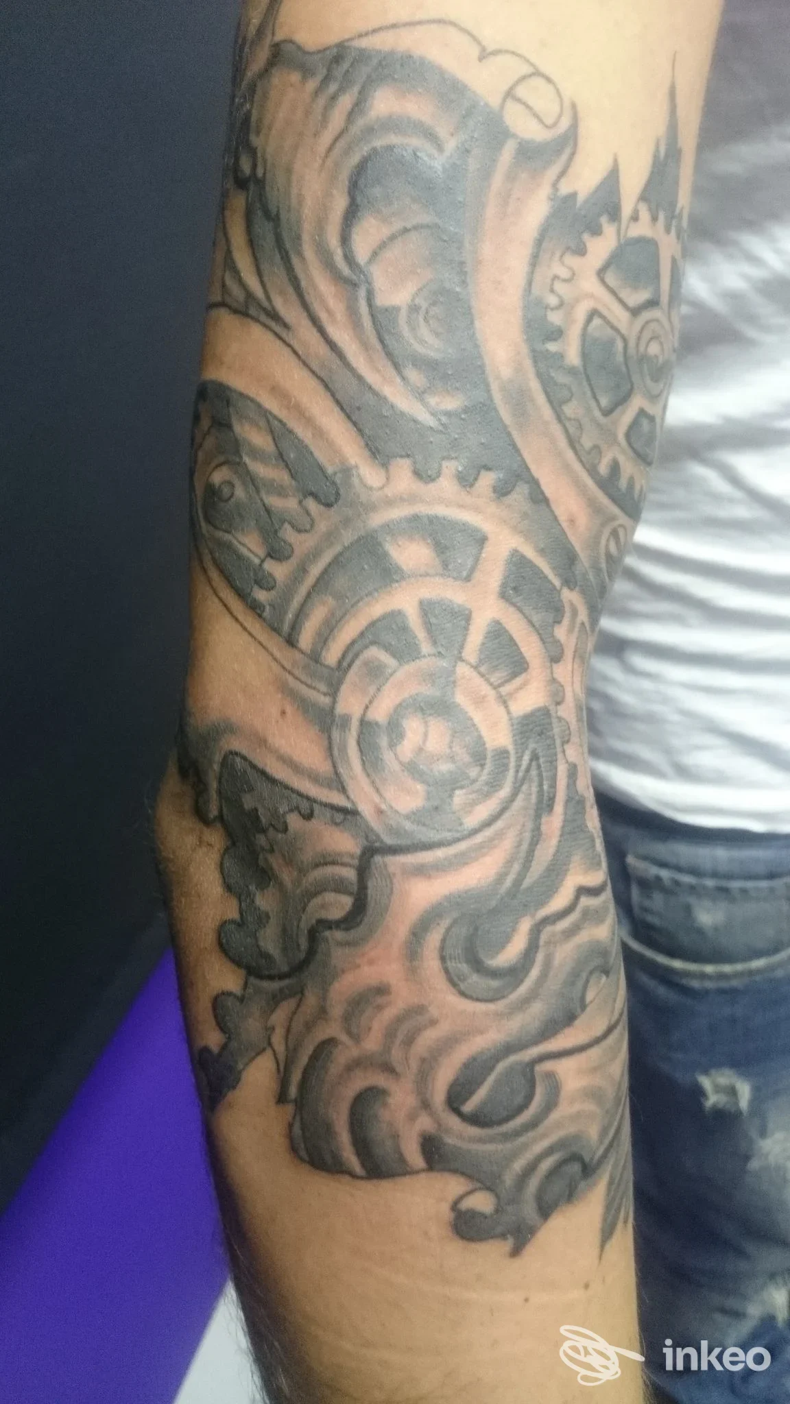 Tattoo visibile su avambraccio con motivi meccanici e ingranaggi, parte di manica visibile; sfondo neutro.