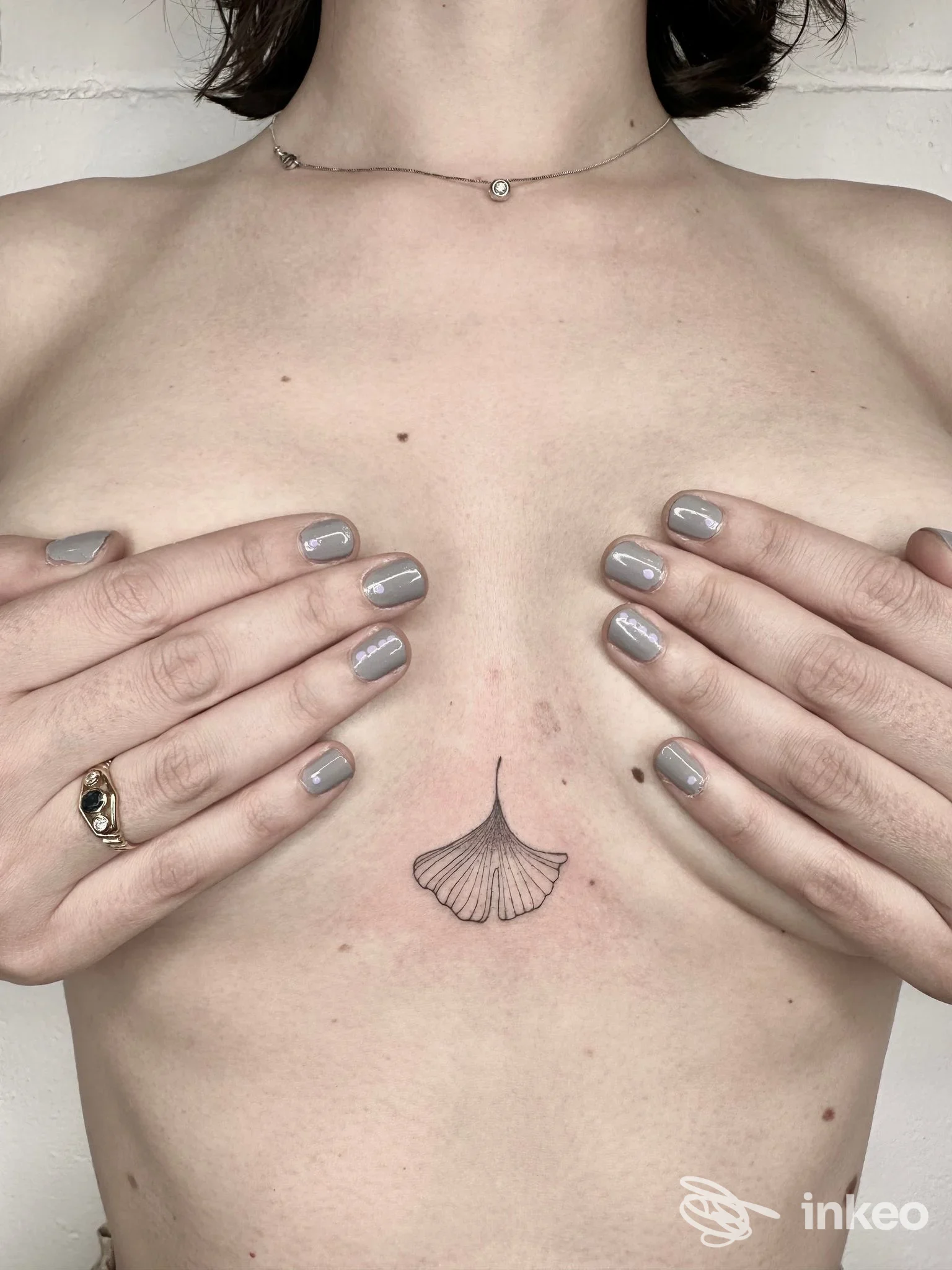 Piccolo tatuaggio a linee sottili a forma di foglia ginkgo sullo sterno, mani che coprono il seno. NEGOZIO-tattoo studio, Gualdo Tadino.