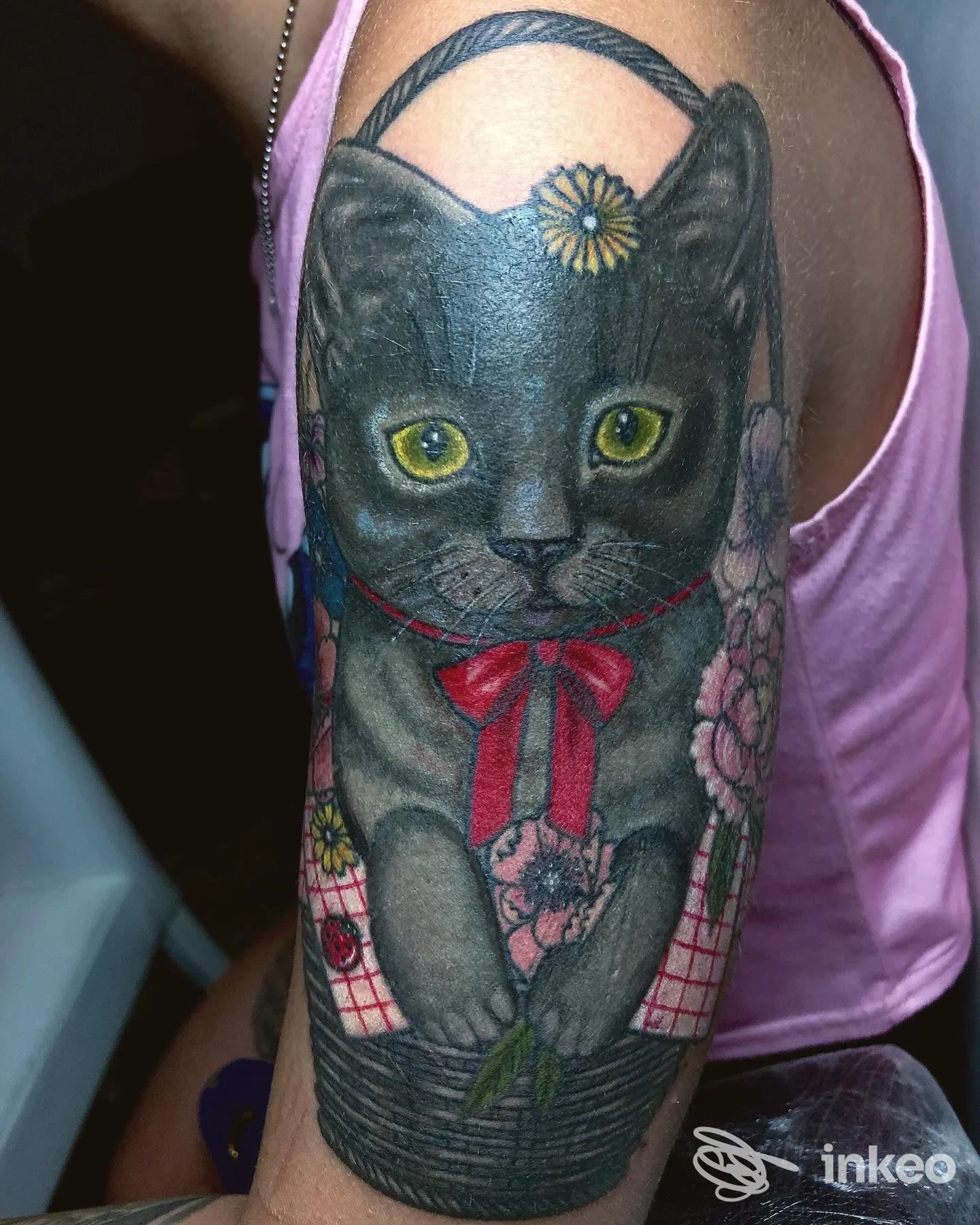 Tatuaggio su braccio superiore raffigurante un gatto nero in un cesto con fiori e nastri rossi. P.Ink Noir Tattoo Studio, Foligno.
