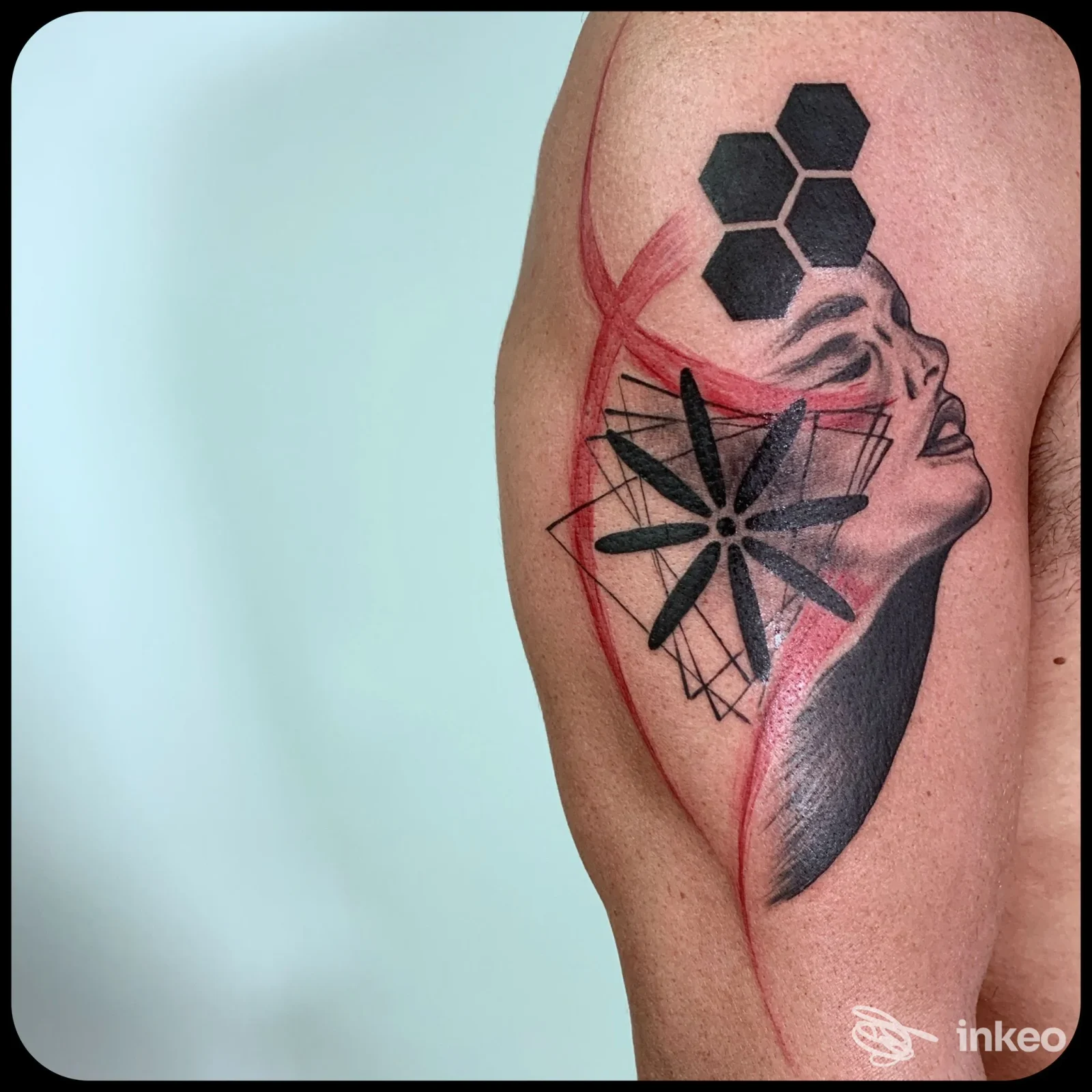 Tatuaggio su braccio superiore: volto stilizzato e elementi geometrici neri con tracce rosse, Punto Tattoo, Terni.