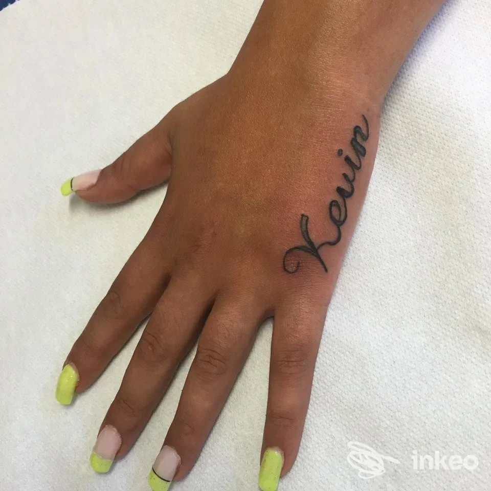 Tatuaggio in corsivo sul dorso della mano, scrittura nera su pelle scura, unghie verniciate in giallo lime, sfondo chiaro.