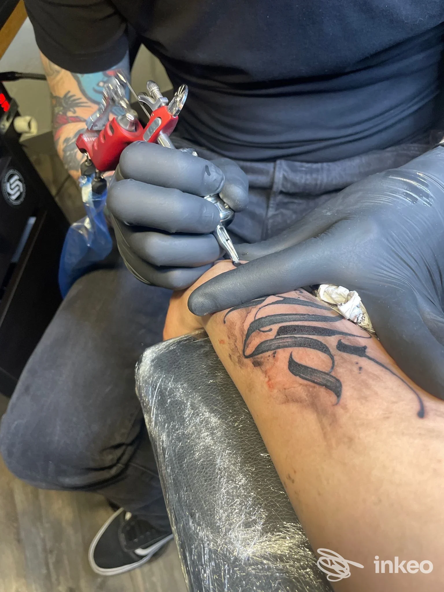 Primo piano di un tatuatore al lavoro su un lettering nero sull'avambraccio del cliente nello studio Jack Puma Tattoo Studio, Aosta.