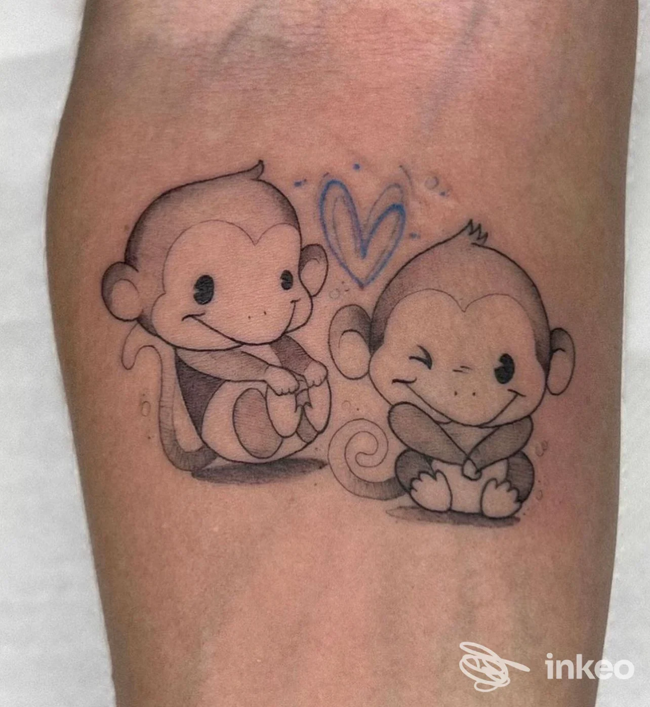 Due tatuaggi raffiguranti due scimmiette stilizzate sedute vicine, una con le mani sulle ginocchia e l'altra che sorride, tra loro un cuore azzurro; tatuaggio su pelle visibile.