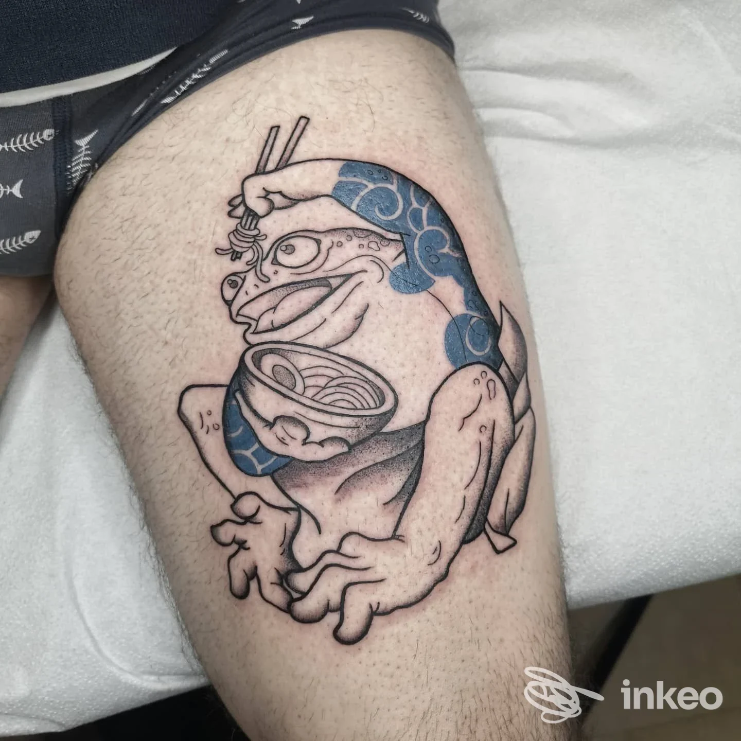 Tatuaggio su coscia raffigurante una rana con ciotola, linee nere e riempiti blu, realizzato da Artium Tattoo Emporium a Montebelluna.