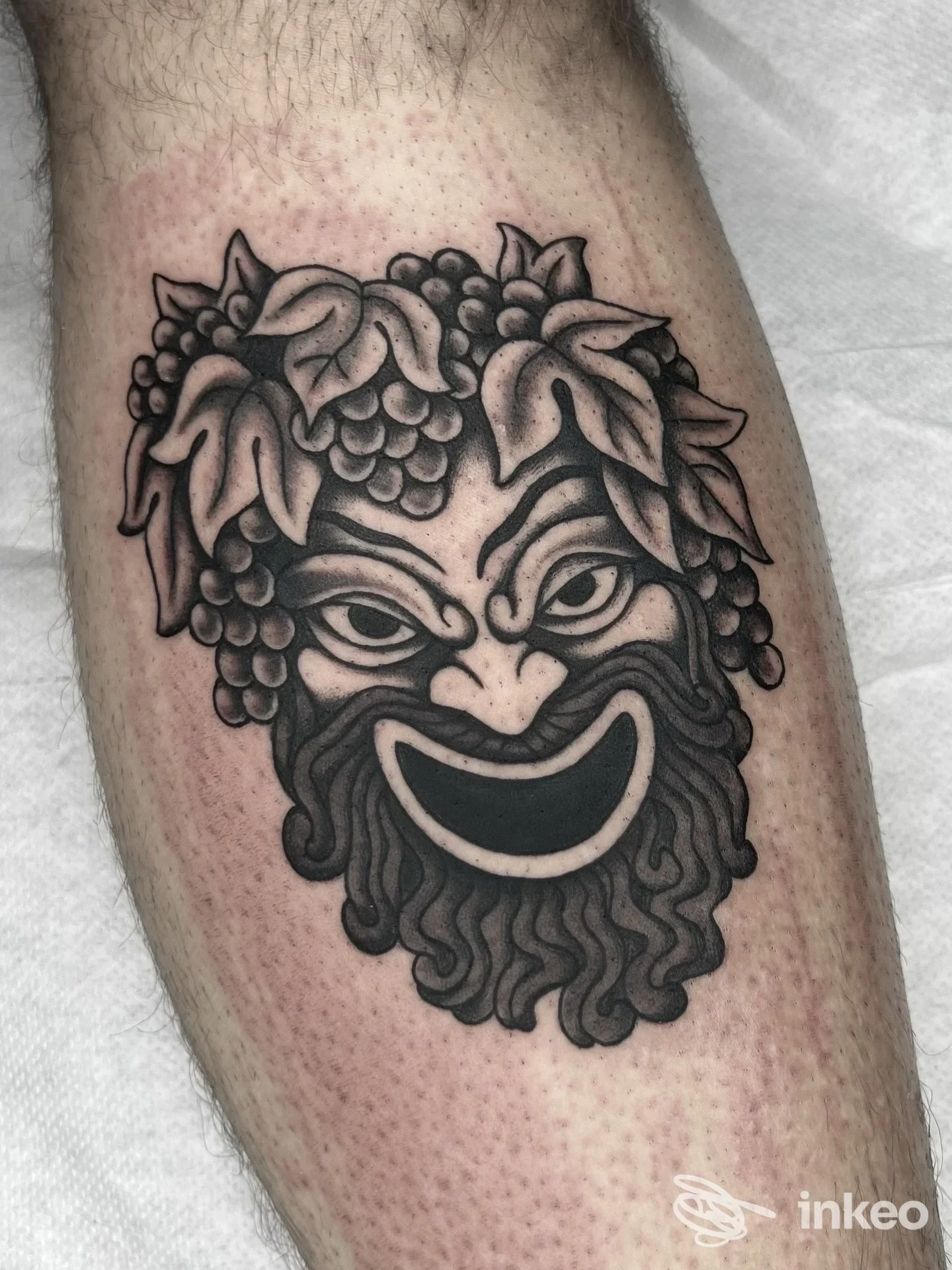 Tatuaggio nero su polpaccio che rappresenta una maschera con foglie e grappoli, foto inviata dallo studio Blk Rooster Tattoo & Piercing, Venezia.
