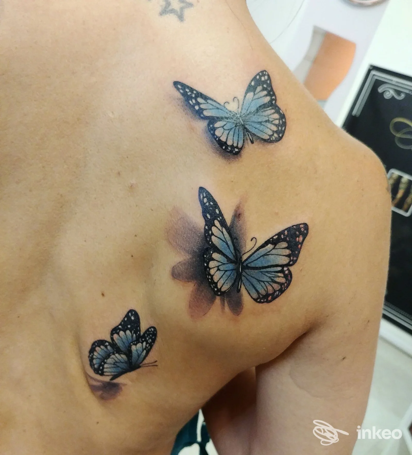 Tre farfalle blu tatuate sulla spalla e sulla parte alta della schiena, con dettagli sottili e sfumature.