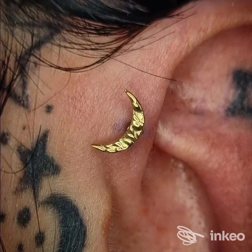 Primo piano di un piercing a mezzaluna dorata sulla conca dell'orecchio, con tatuaggi vicini. GioRGia BodyPiercer, San Donà di Piave.