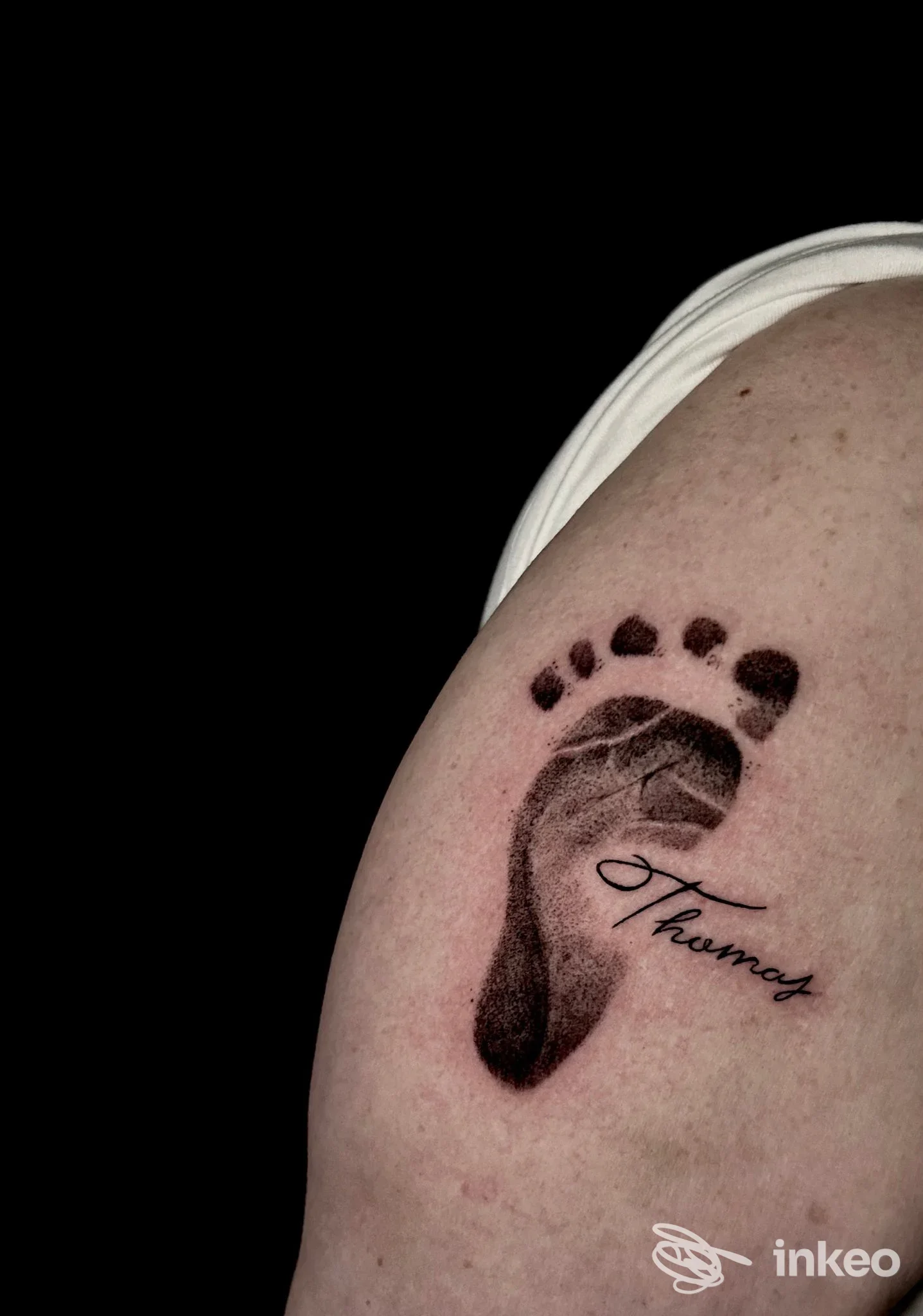 Un tatuaggio su pelle visibile raffigura un piccolo piede con dita e una scritta elegante Thomas, situato sull’avambraccio/sovracciglio della spalla, su sfondo nero.